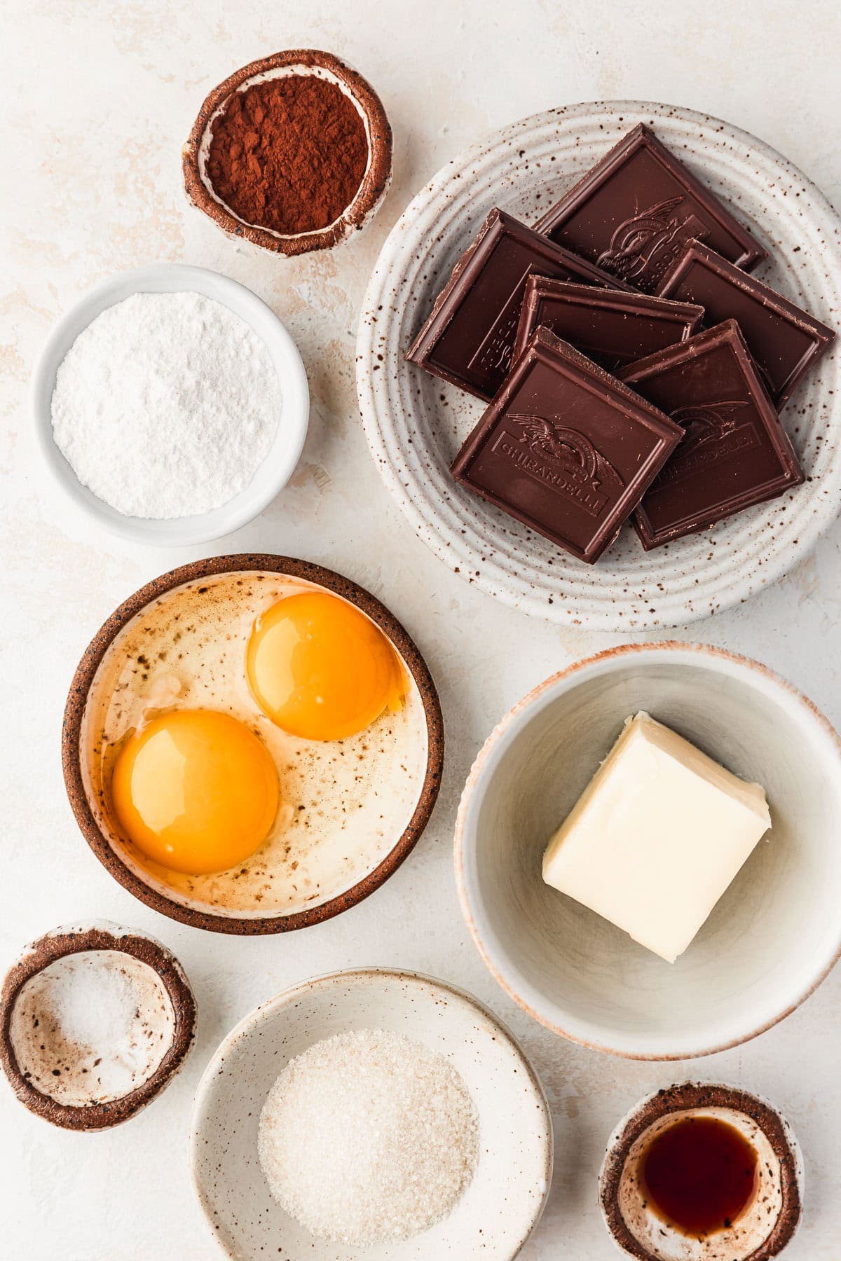 White and brown bowls of chocolate, cocoa powder, flour, butter, eggs, sugar, salt, and vanilla on a tan counter.