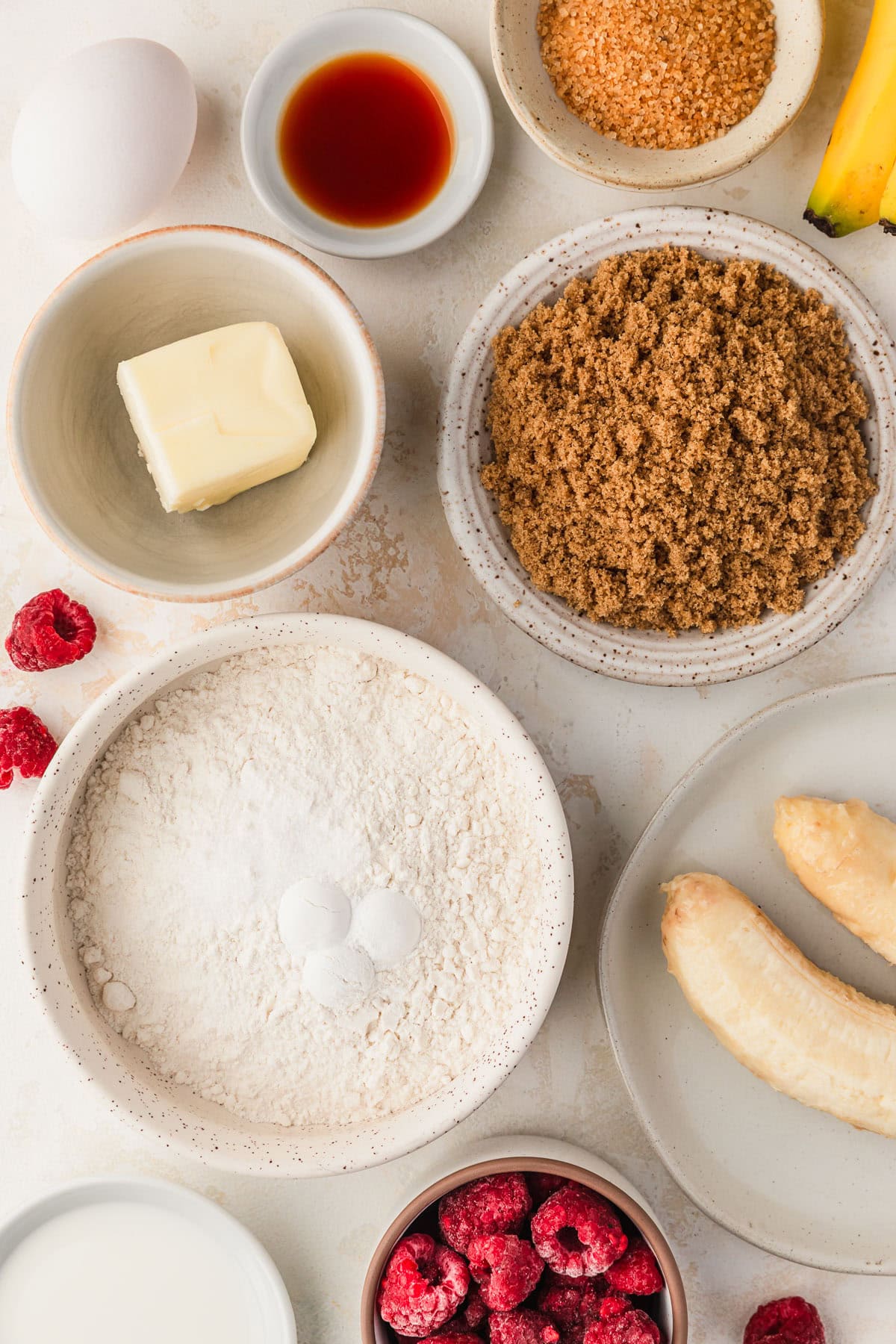 White bowls of brown sugar, flour, butter, bananas, raspberries, raw sugar, vanilla, and milk next to an egg on a beige table.