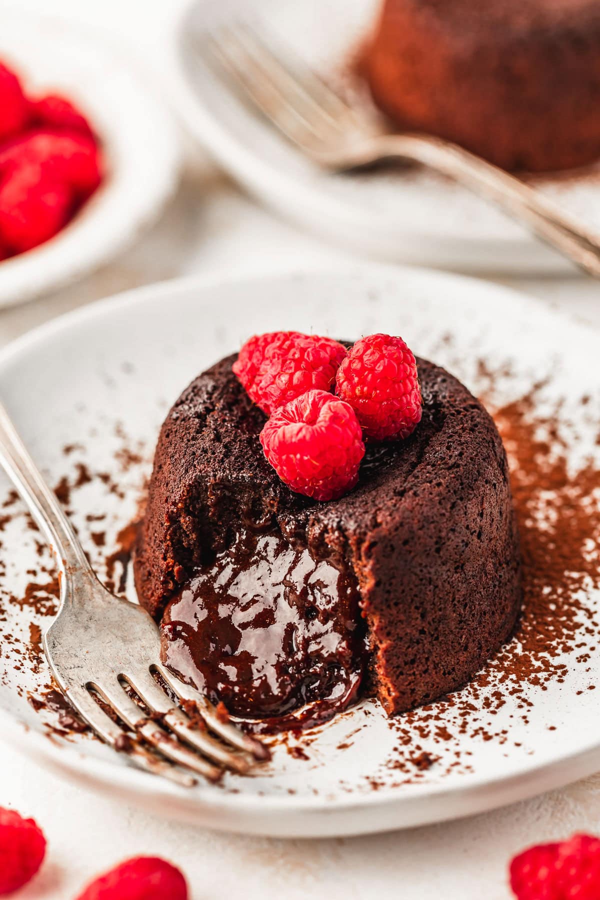 Two white plates with molten lava cakes, berries, and forks and a white bowl of raspberries on a white counter.