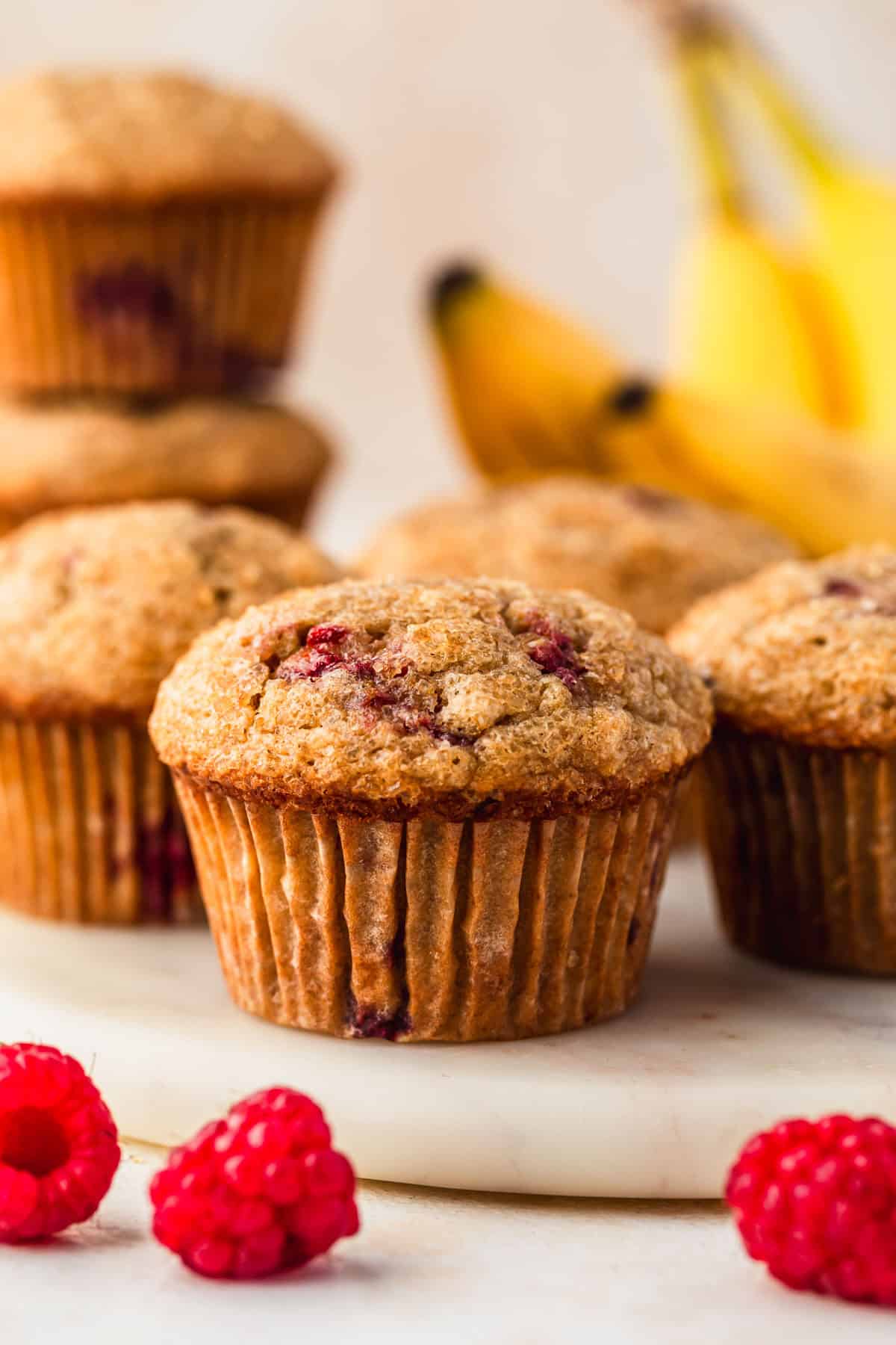 A white tray of small batch raspberry banana muffins next to a bunch of bananas and raspberries on a tan counter.