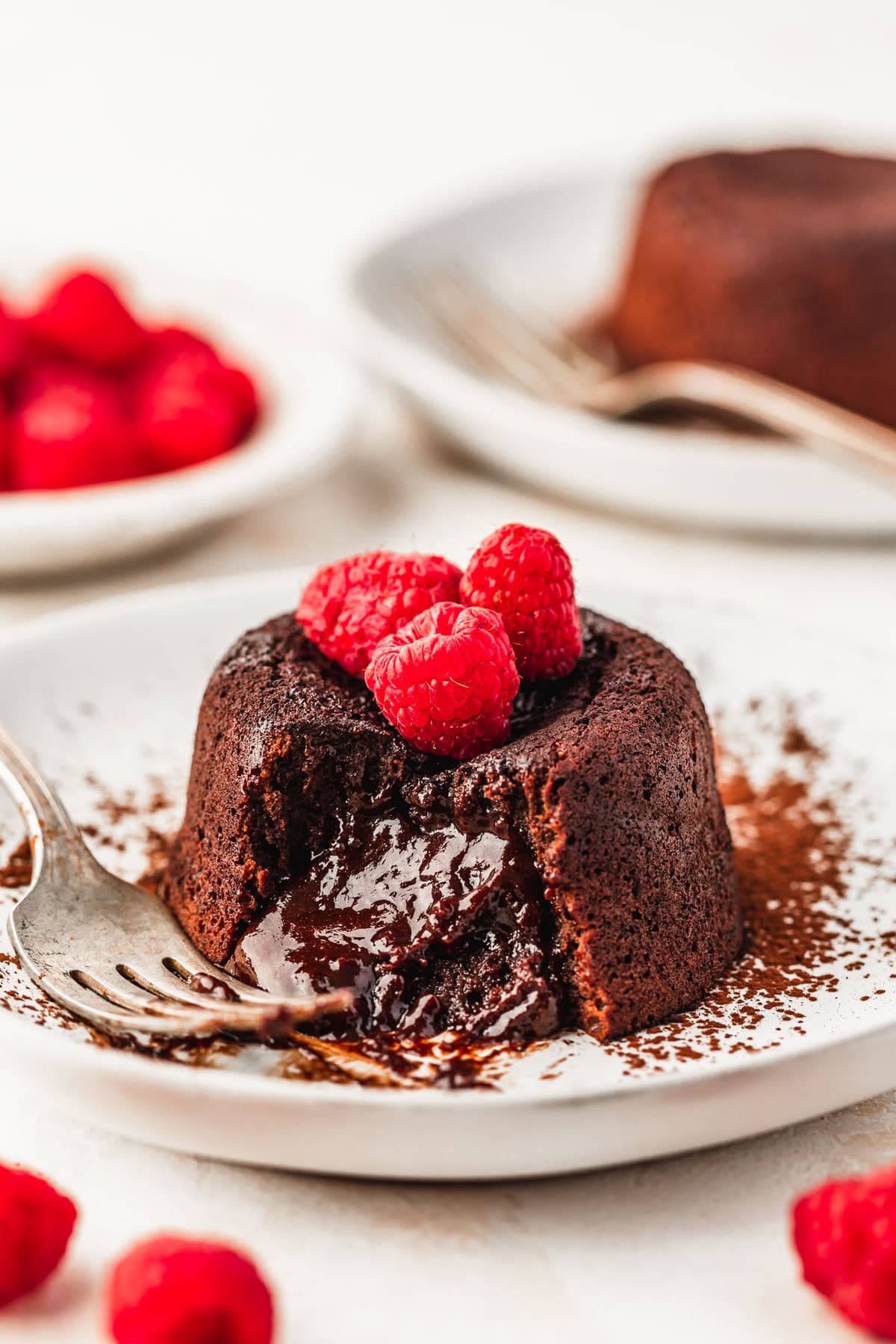 Two white plates with molten lava cakes, berries, and forks next to a white bowl of raspberries with a white background.