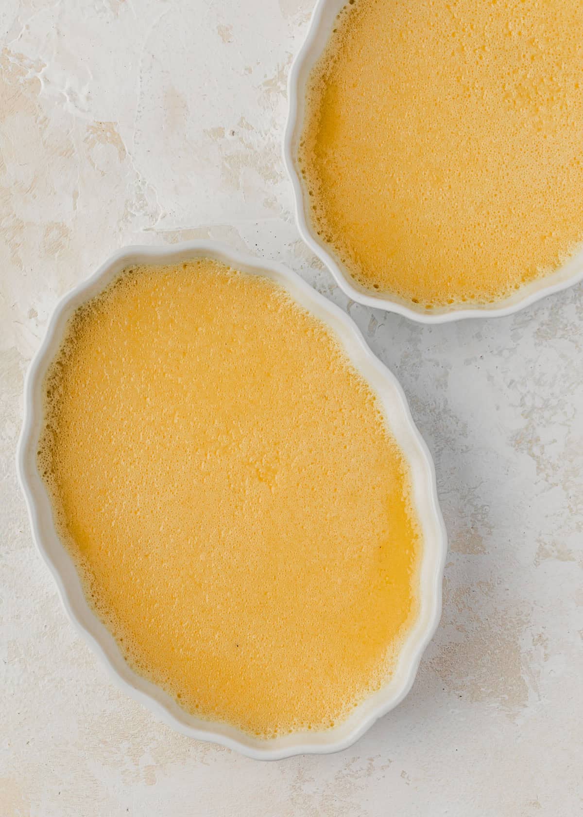 Two ramekins of crème brûlée custard on a white counter.