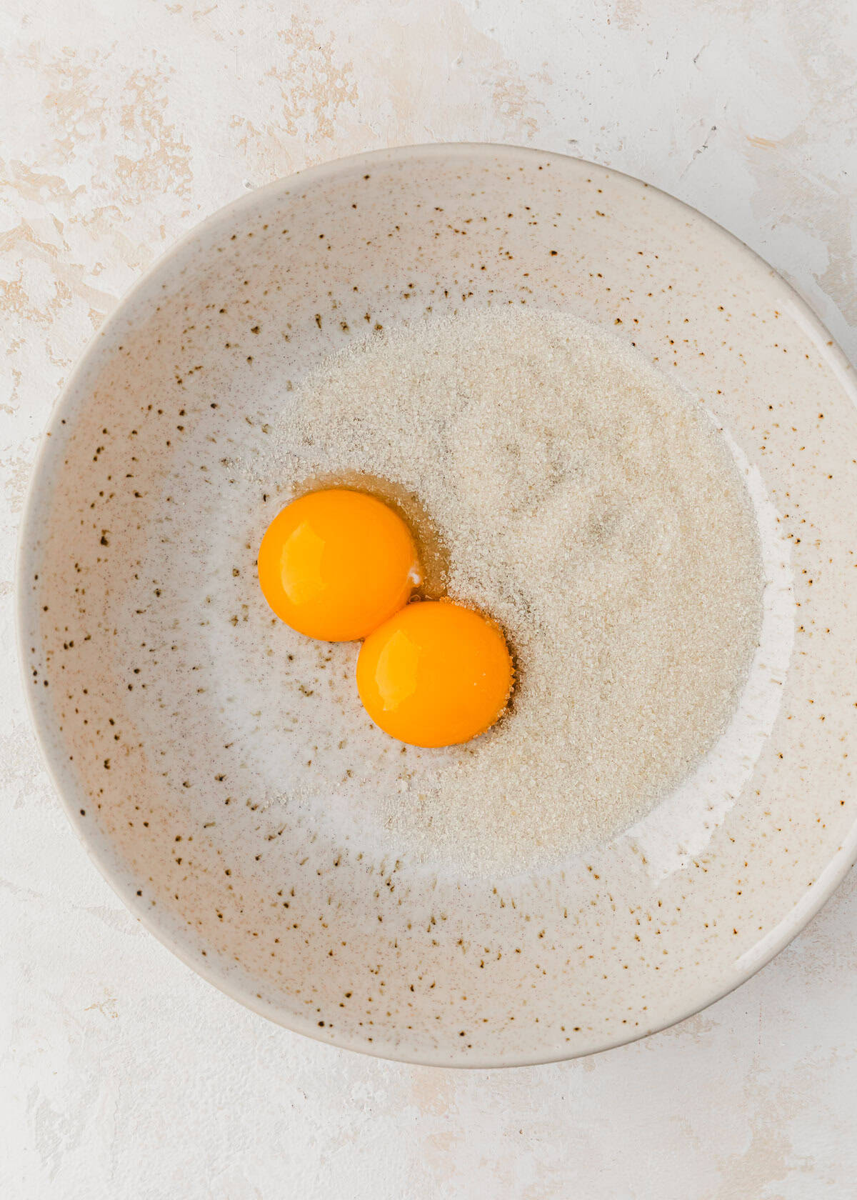A white bowl of sugar and egg yolks on a white counter.