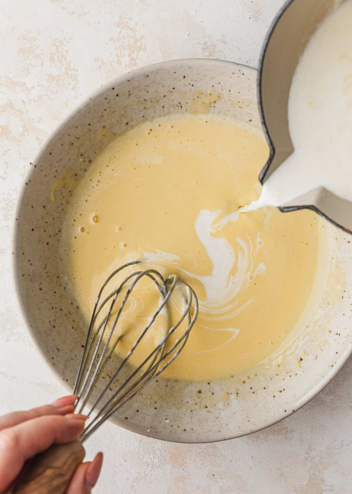 A hand pouring a pot of cream into a white bowl of egg yolks and sugar.