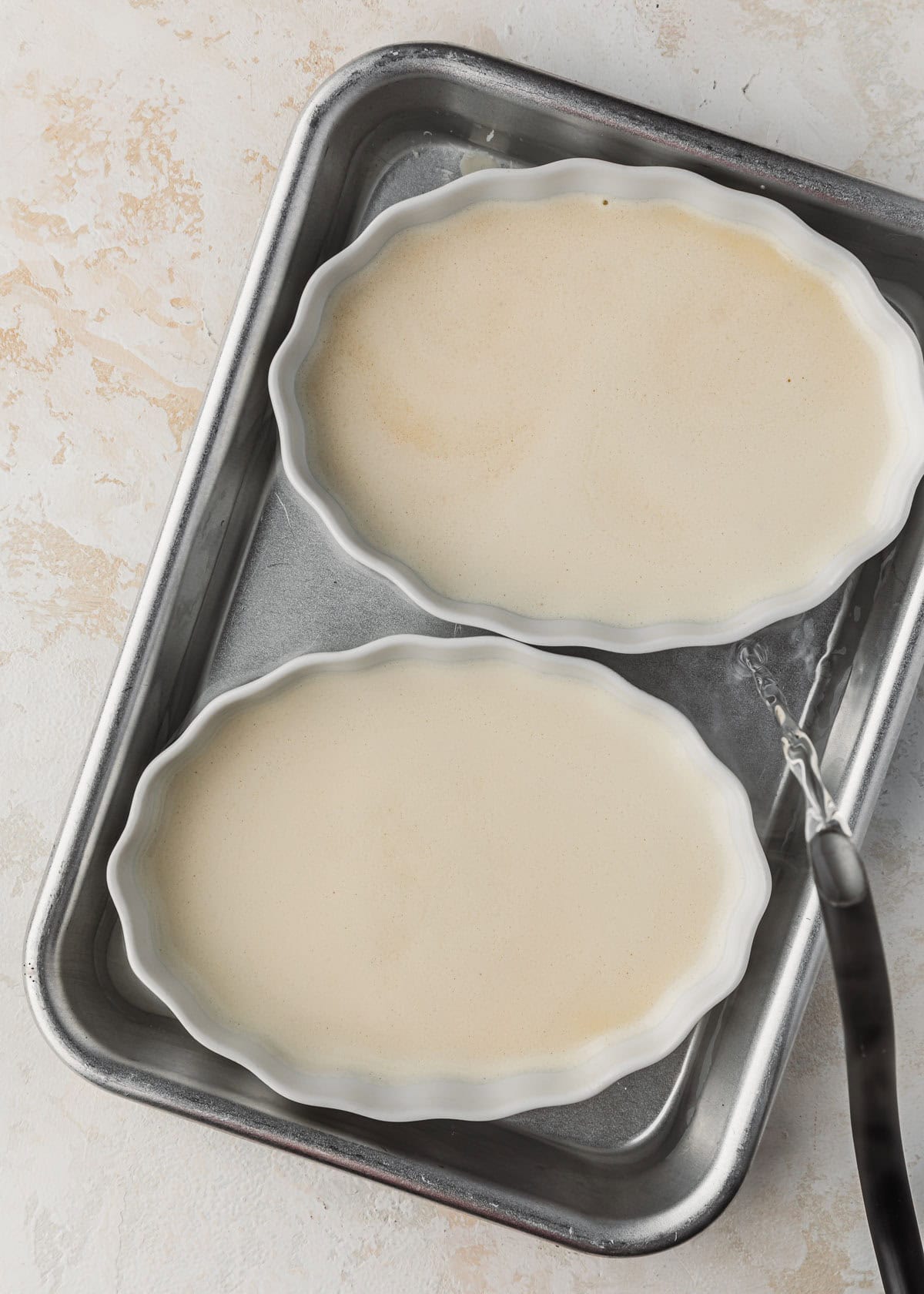 A kettle pouring water into a pan with two ramekins of crème brûlée.