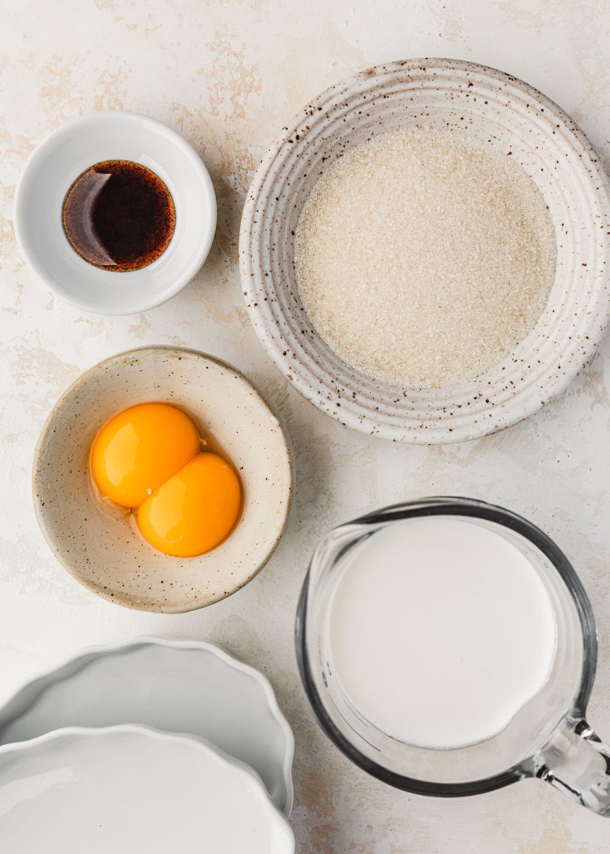 White bowls of sugar, egg yolks, and vanilla bean paste on a white counter next to a pitcher of cream and white ramekins.