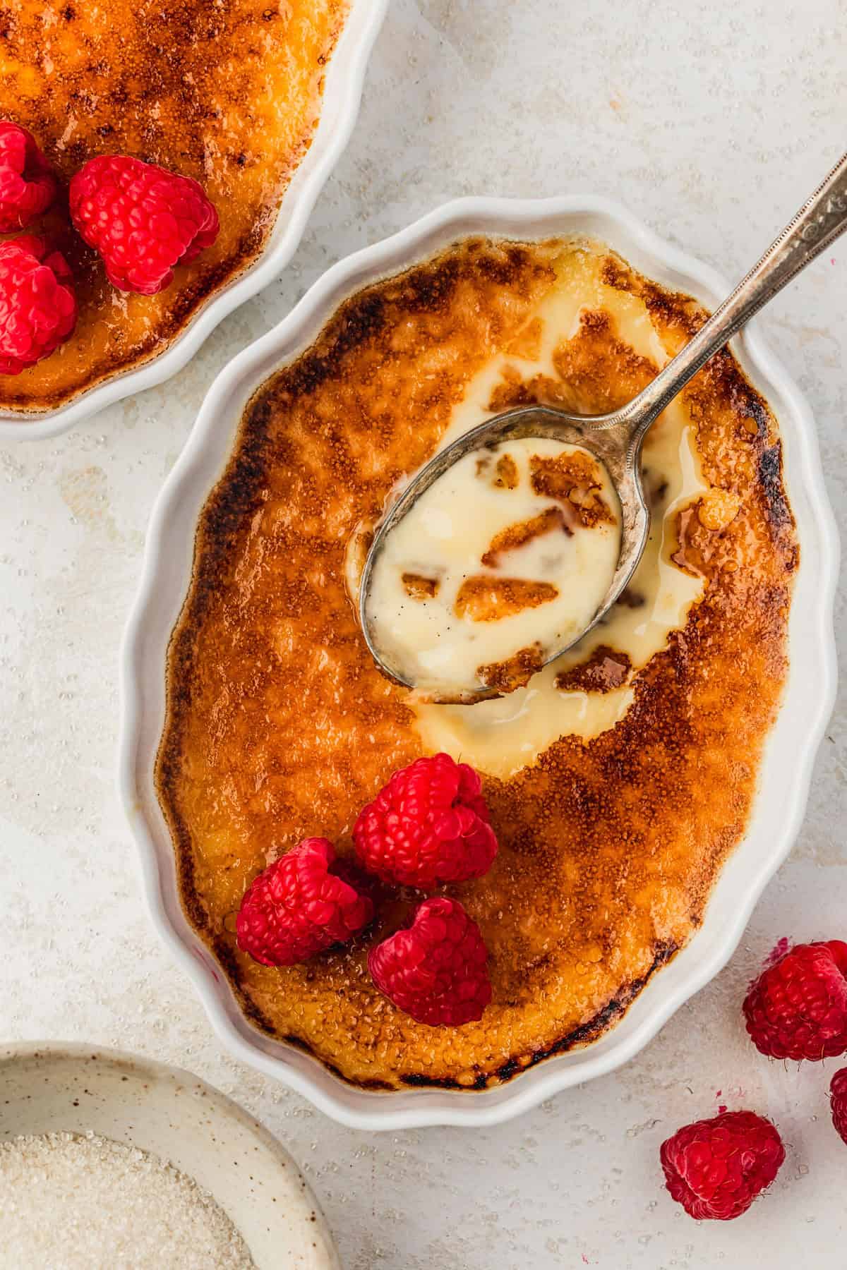 Two ramekins of crème brûlée with raspberries, one with a spoon and cracked sugar, next to more berries and a white bowl of sugar with a white and beige speckled background.