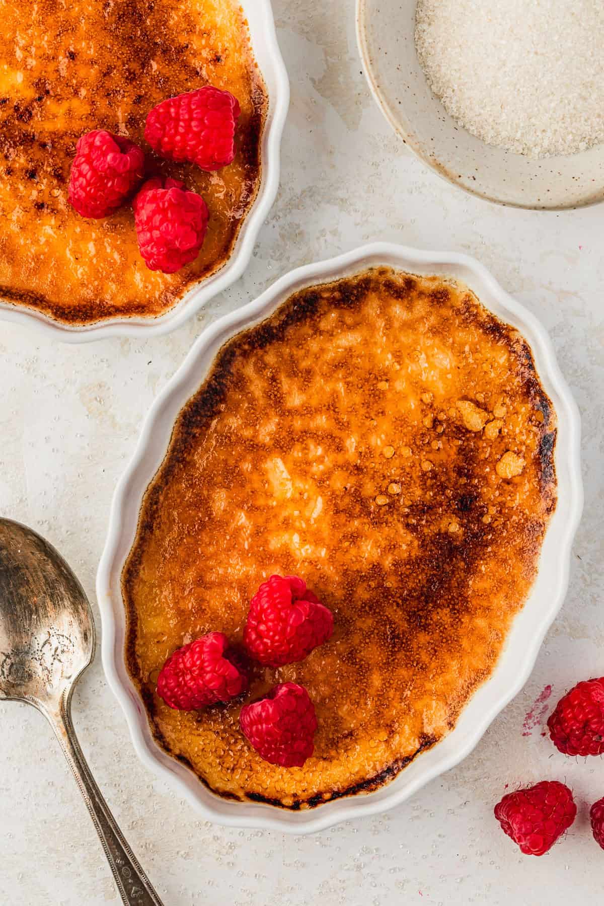 Two ramekins of crème brûlée with raspberries on a white counter next to a spoon and white bowl of sugar.