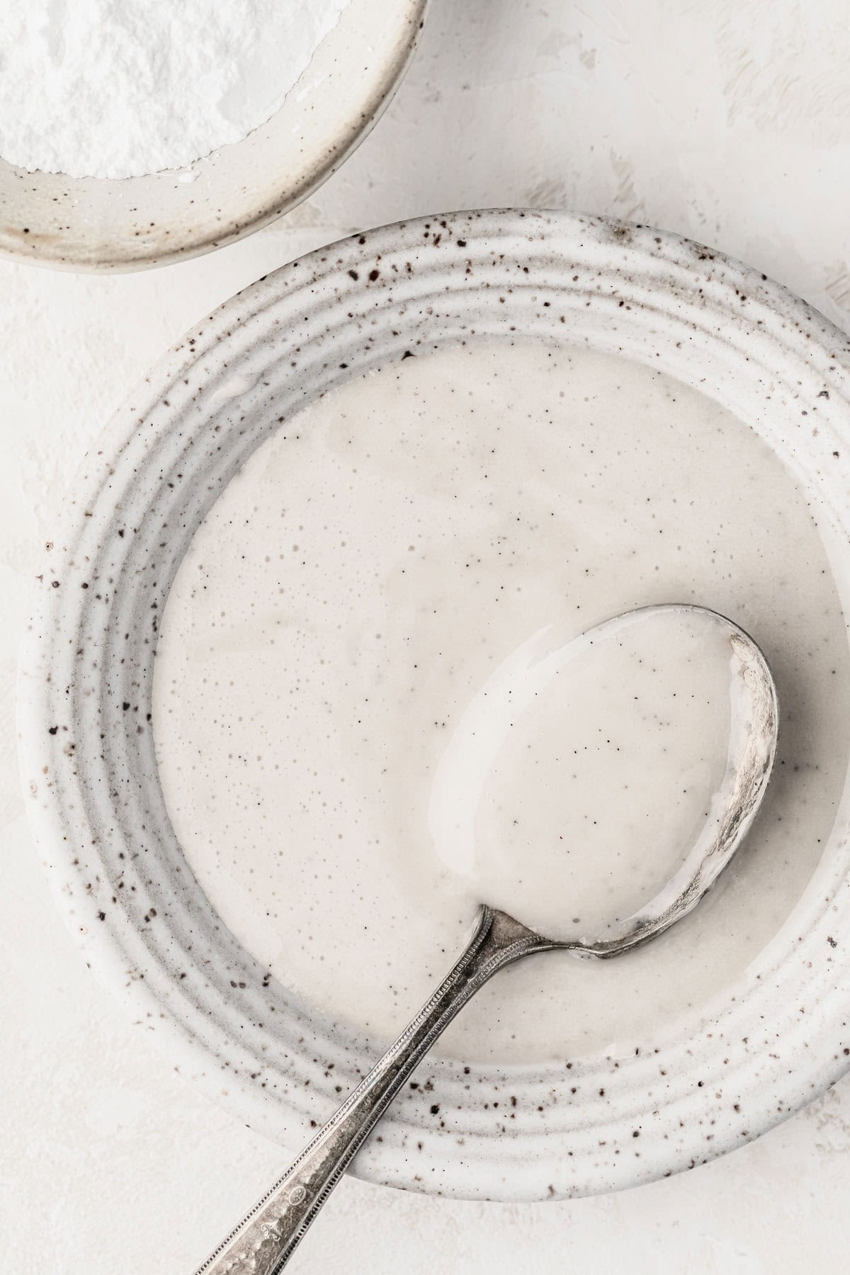 A white bowl of small batch vanilla bean glaze with a spoon on a white counter next to a white bowl of powdered sugar.