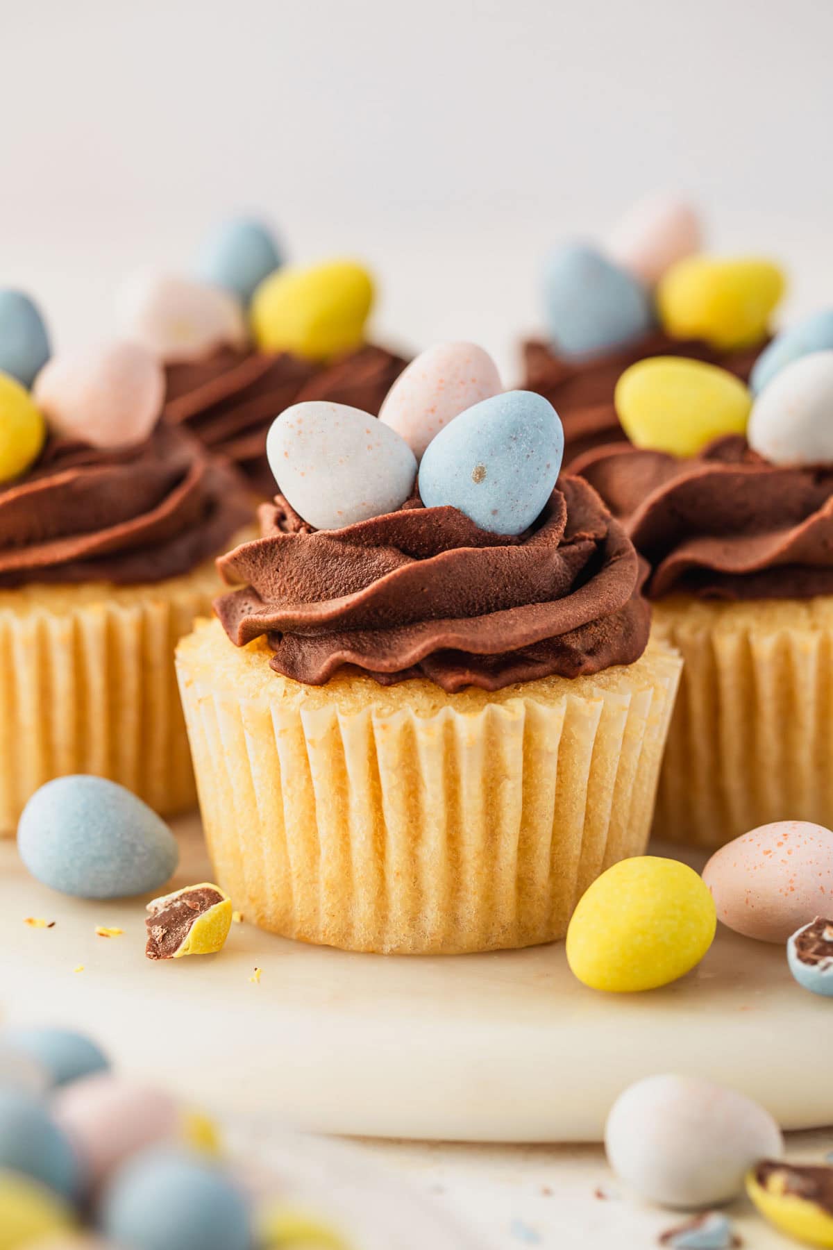 A white marble platter of small batch Easter egg cupcakes with mini egg candies next to a white bowl of candies on a white background.