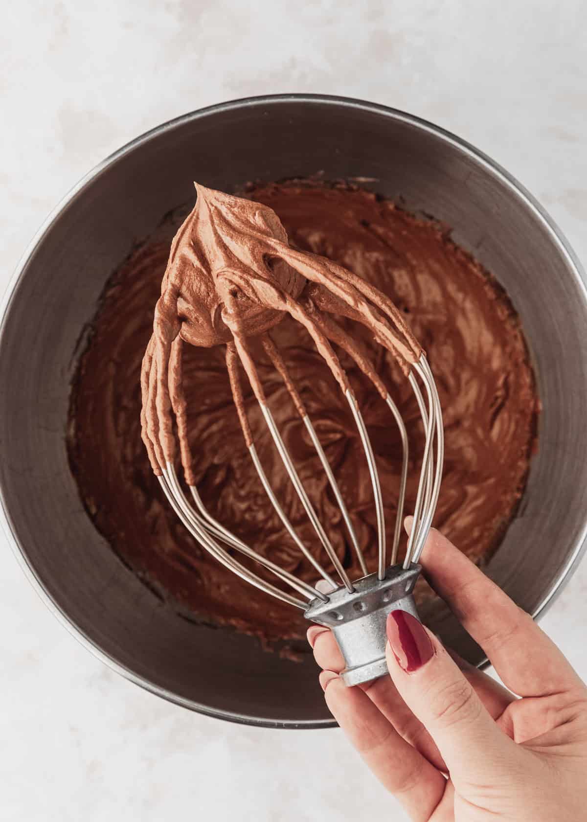 A hand holding a whip attachment over a bowl of chocolate buttercream for small batch Easter egg cupcakes.