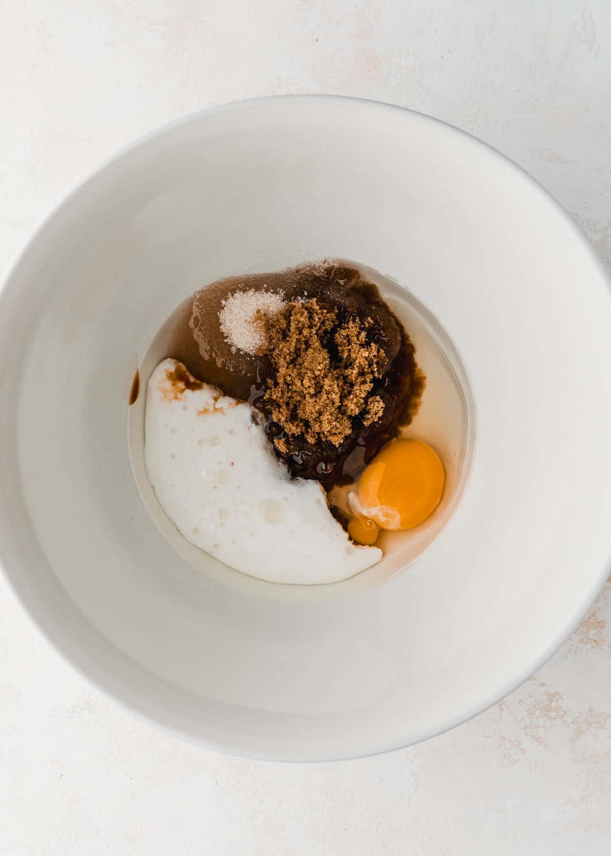 A white bowl of oil, sugar, vanilla, and an egg on a white counter.