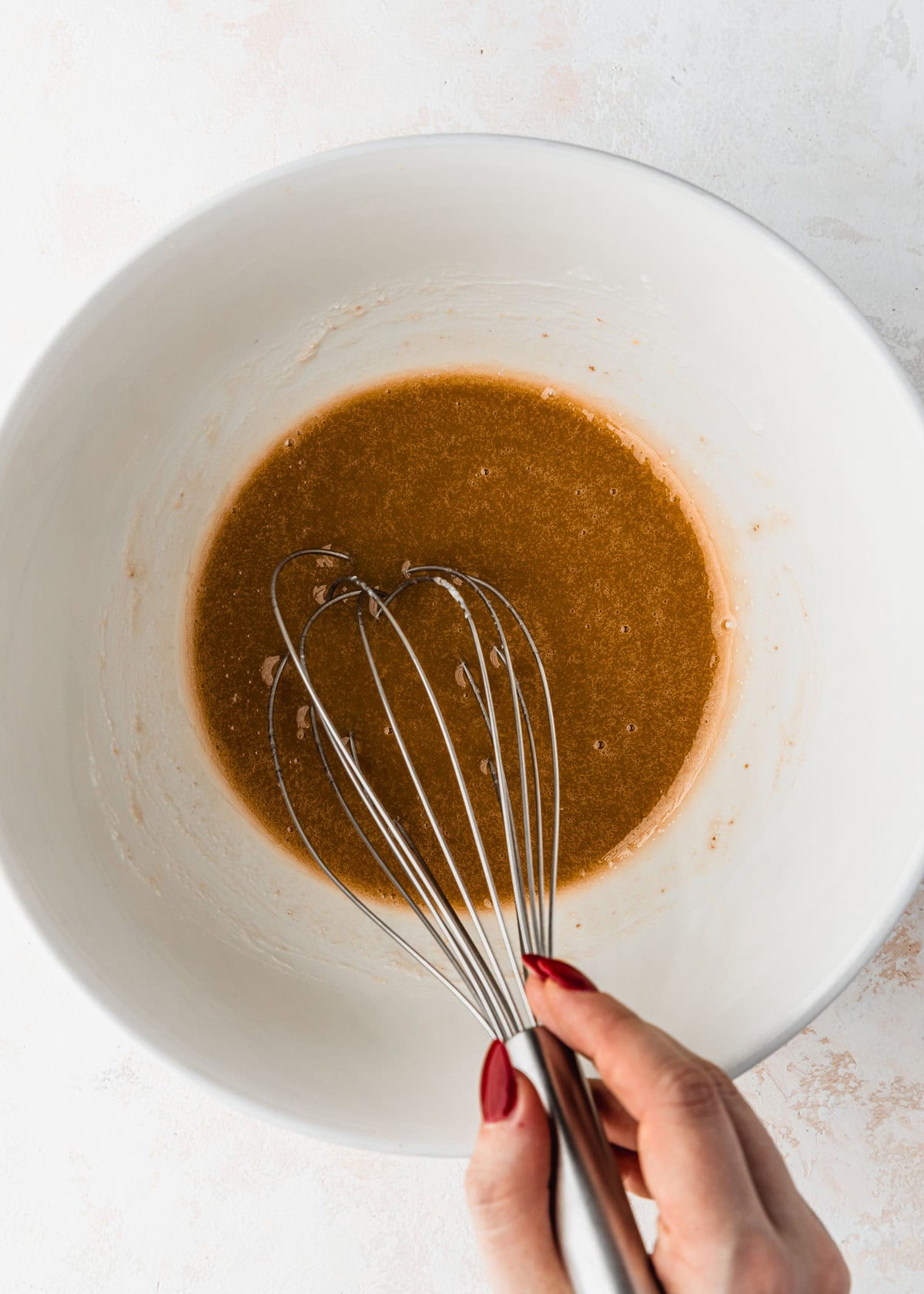 A hand using a whisk to mix oil, sugar, and an egg in a white bowl.