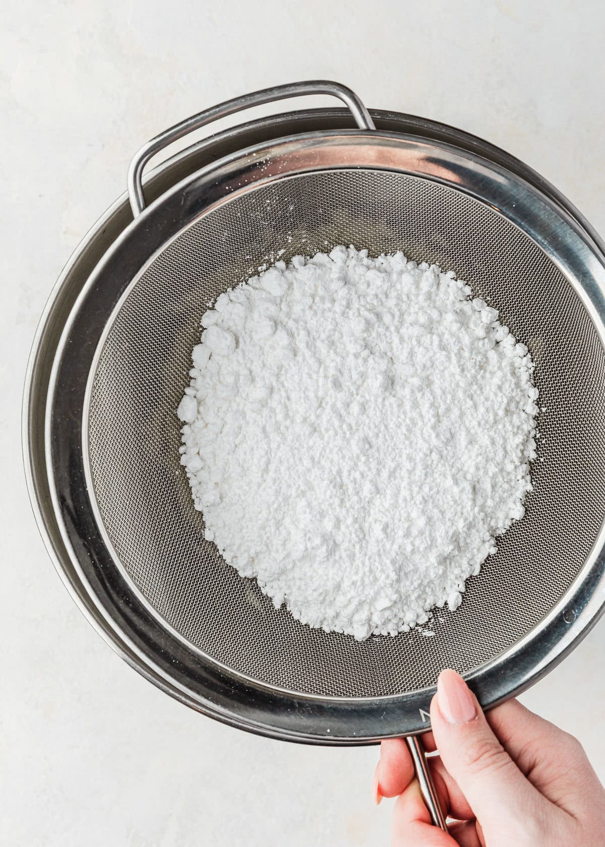 A hand using a sieve to sift powdered sugar into a silver bowl.