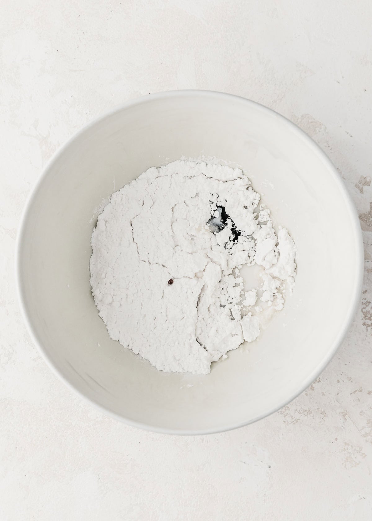 A white bowl of powdered sugar, milk, and vanilla bean paste on a white counter.