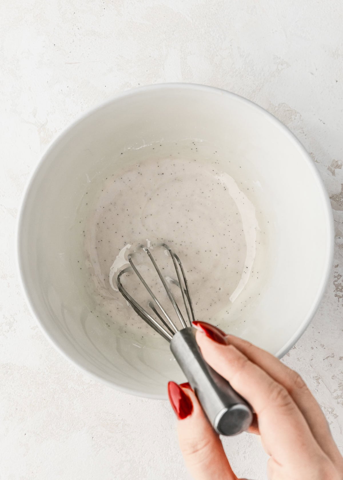 A hand using a whisk to mix vanilla icing in a white bowl.