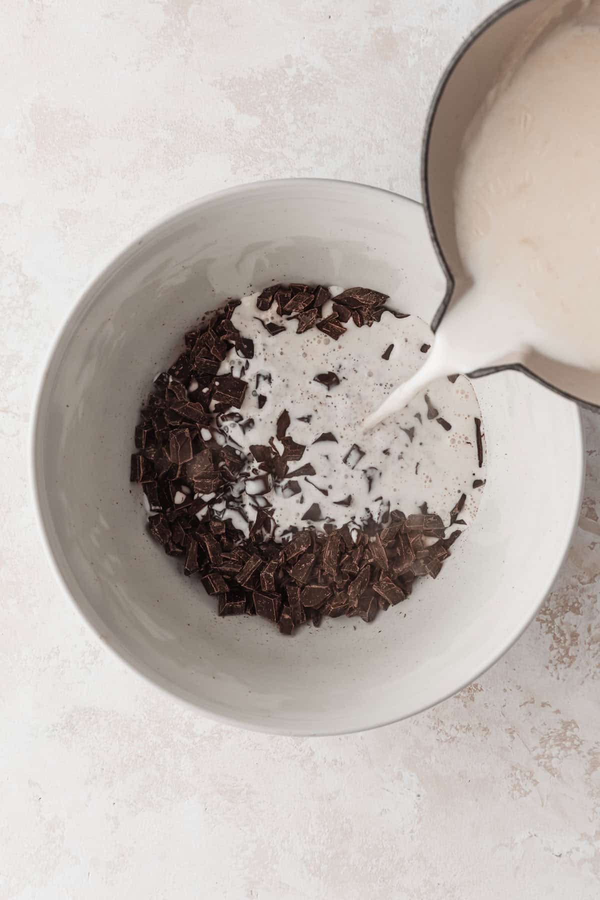 A hand pouring a pot of hot cream over a white bowl of chopped chocolate. 