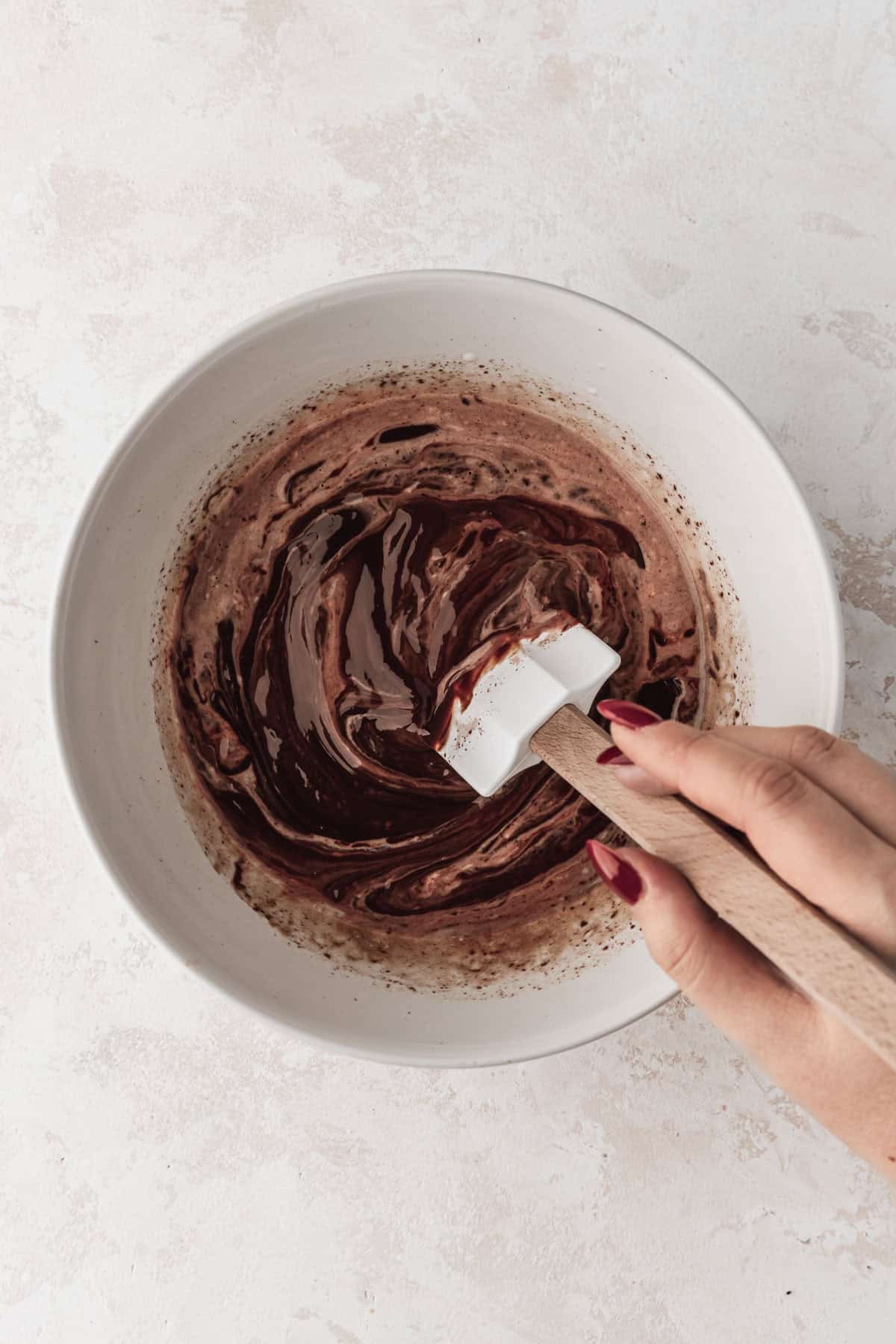 A hand using a rubber spatula to stir ganache in a white bowl.