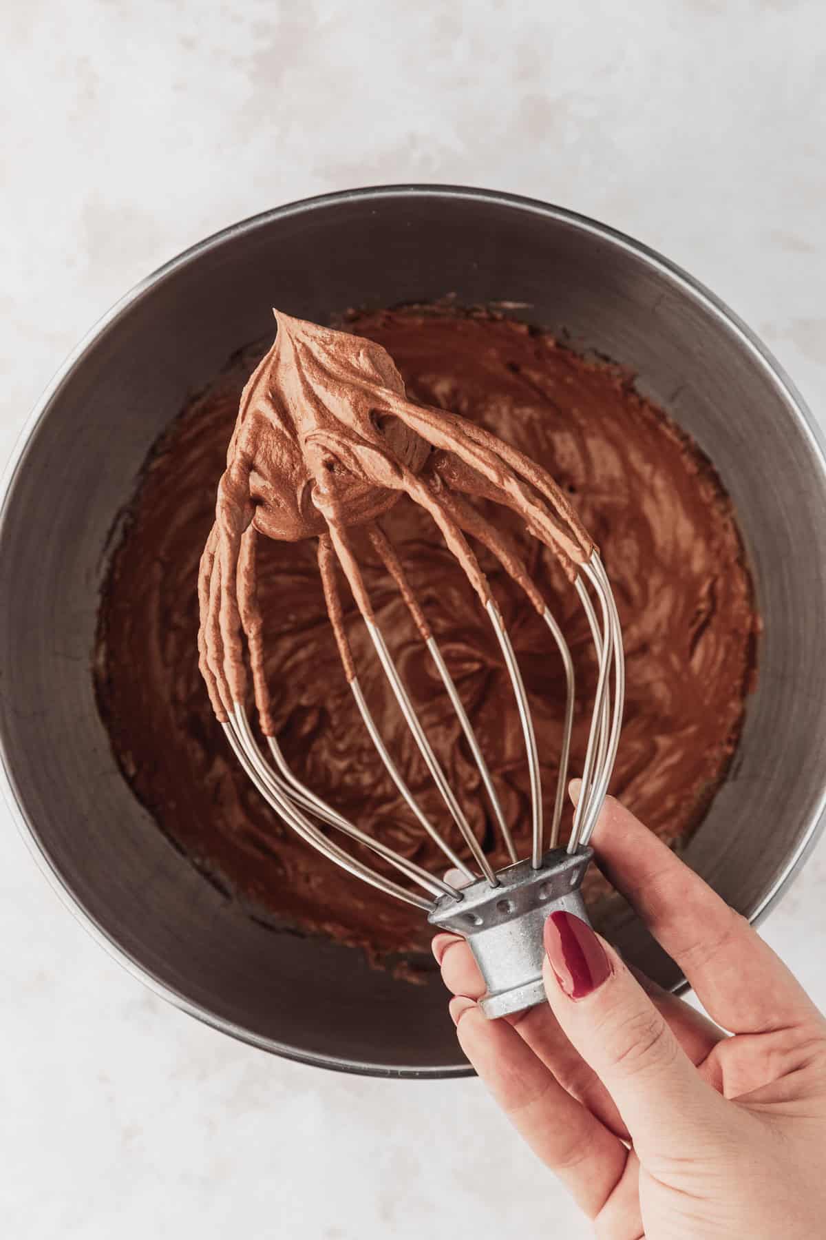 A hand holding a whip attachment over a silver bowl of whipped chocolate ganache.