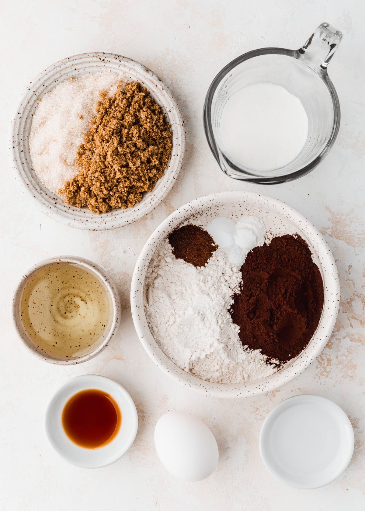 White bowls of flour, cocoa powder, oil, sugar, brown sugar, buttermilk, water, and vanilla next to an egg on a tan counter.
