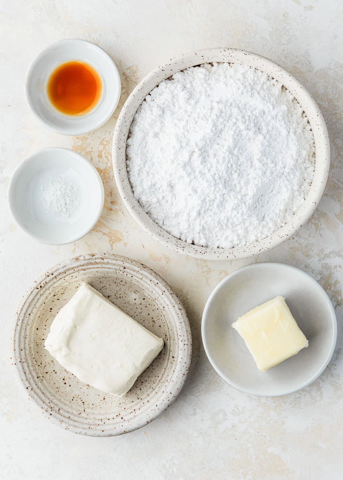 White bowls of powdered sugar, vanilla, salt, cream cheese, and butter on a tan counter.