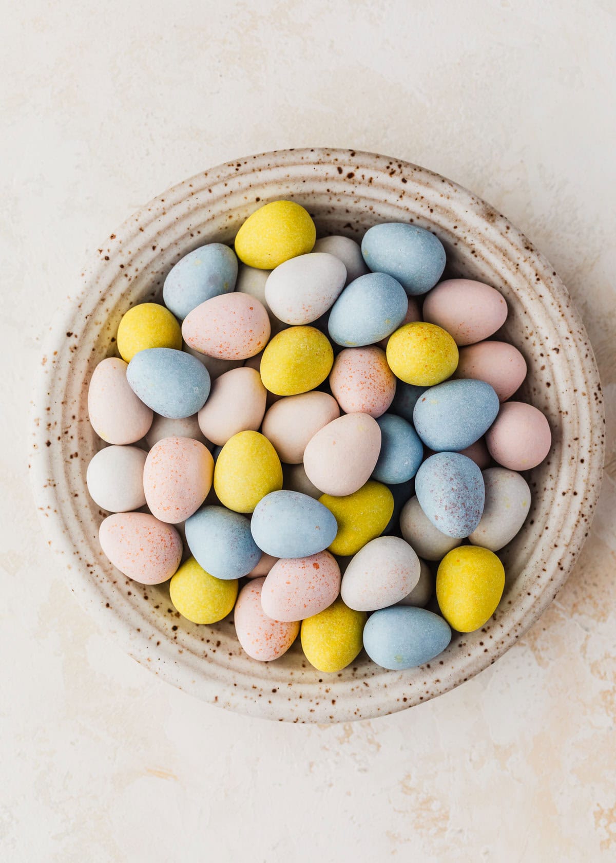A white bowl of Cadbury mini egg candies on a tan counter.