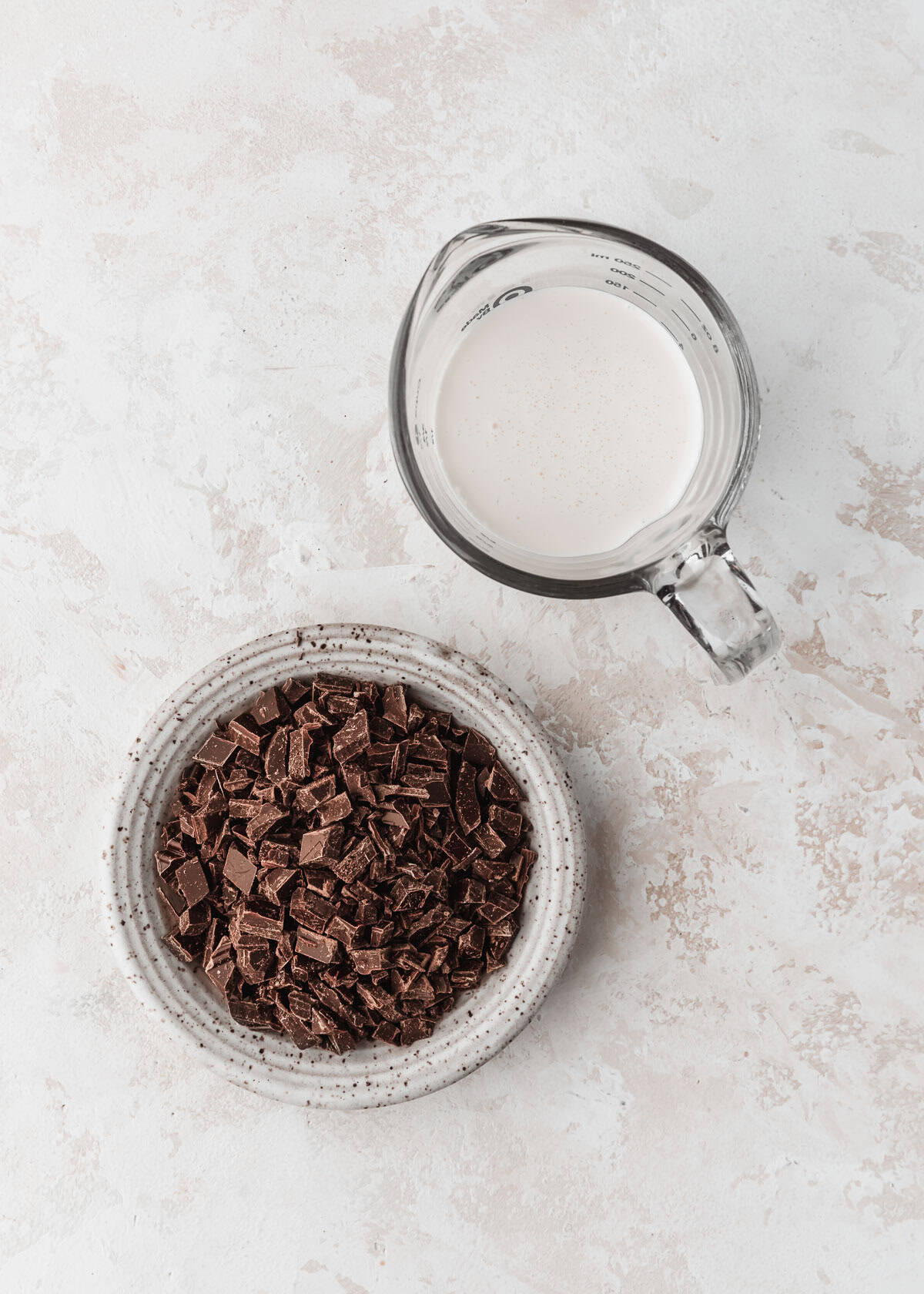 A white bowl of chocolate next to a pitcher of heavy cream on a white counter.