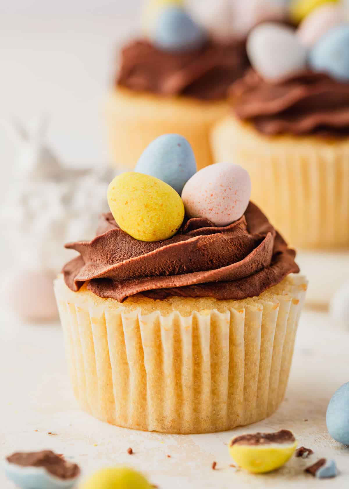 A cupcake next to ceramic Easter bunnies, egg candies, and a white platter of small batch Easter egg cupcakes on a white background.