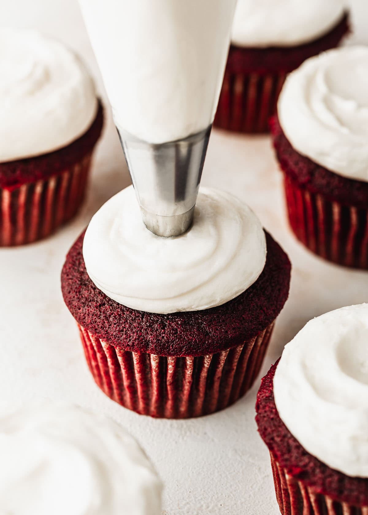A piping bag piping small batch cream cheese frosting on a cupcake next to rows of red velvet cupcakes on a white counter.