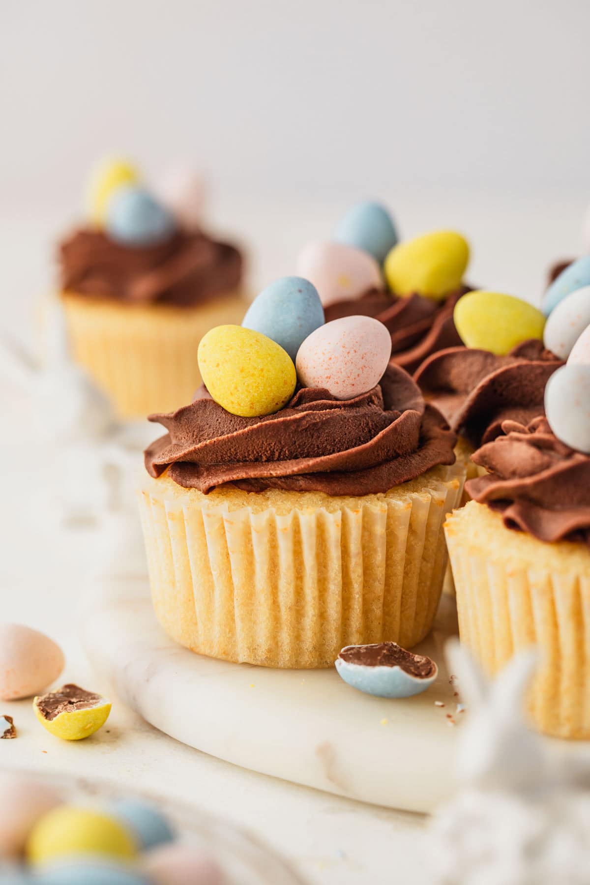 A white platter of small batch Easter egg cupcakes with Cadbury mini eggs on a white counter next to ceramic bunnies and a white bowl of mini egg candies.
