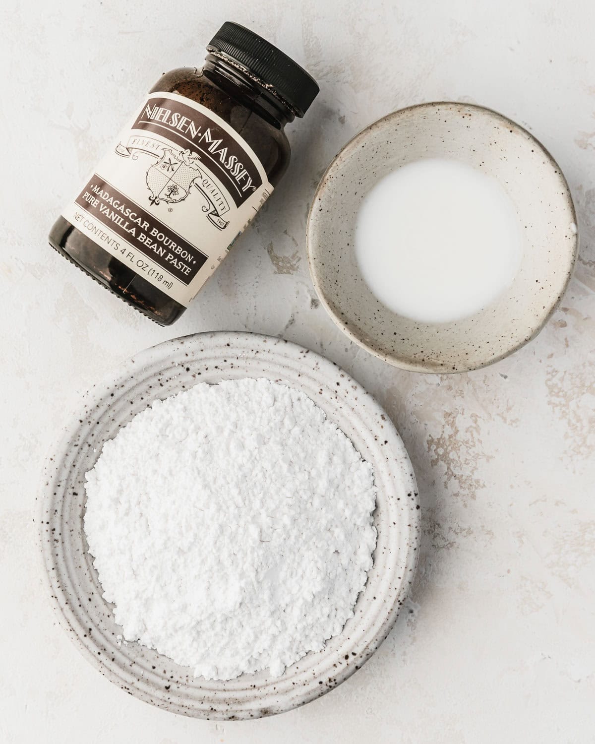 White bowls of milk and powdered sugar next to a jar of vanilla bean paste on a white counter.
