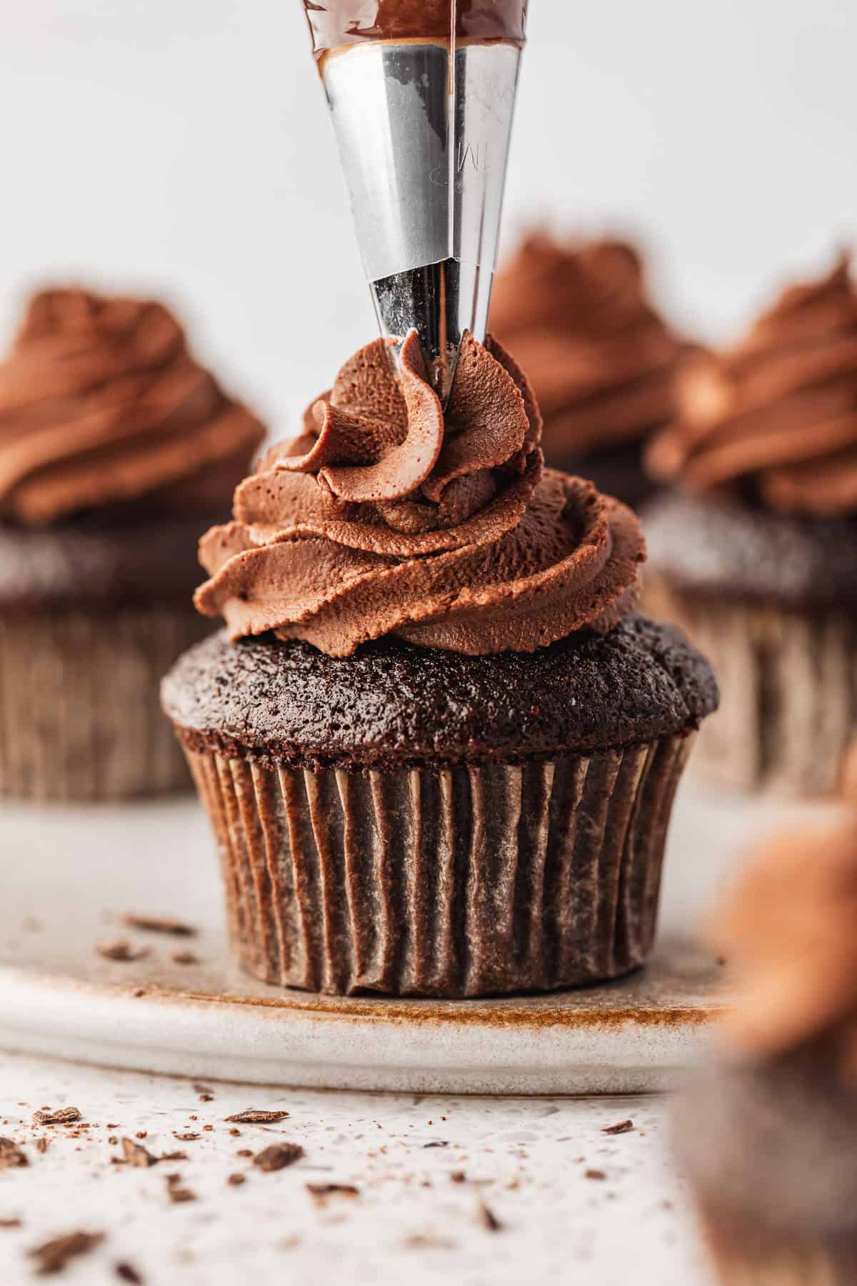 A piping bag decorating a cupcake with whipped chocolate ganache. The cupcake is on a grey platter next to more cupcakes.
