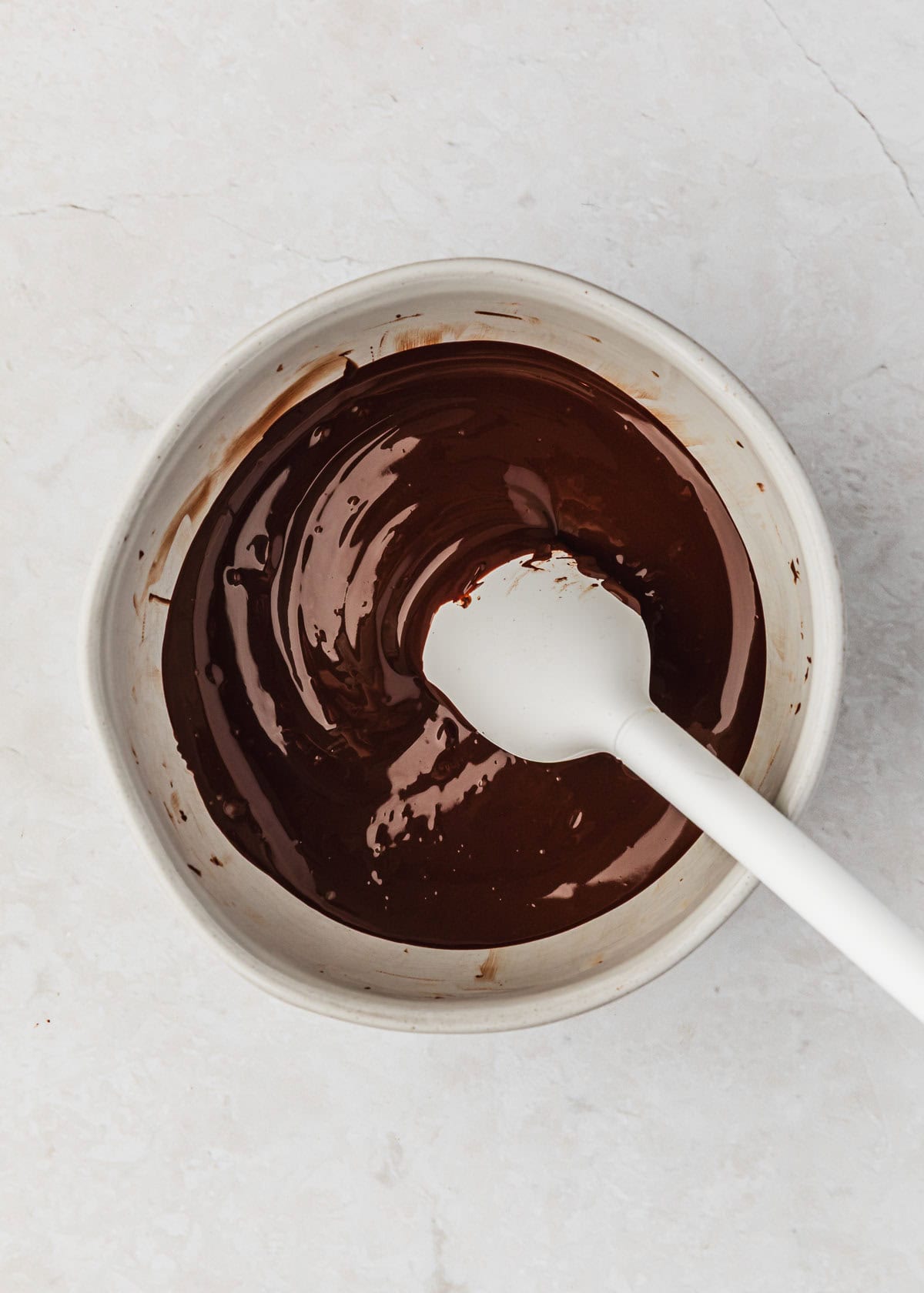 A white bowl of melted chocolate with a white rubber spatula on a marble counter.