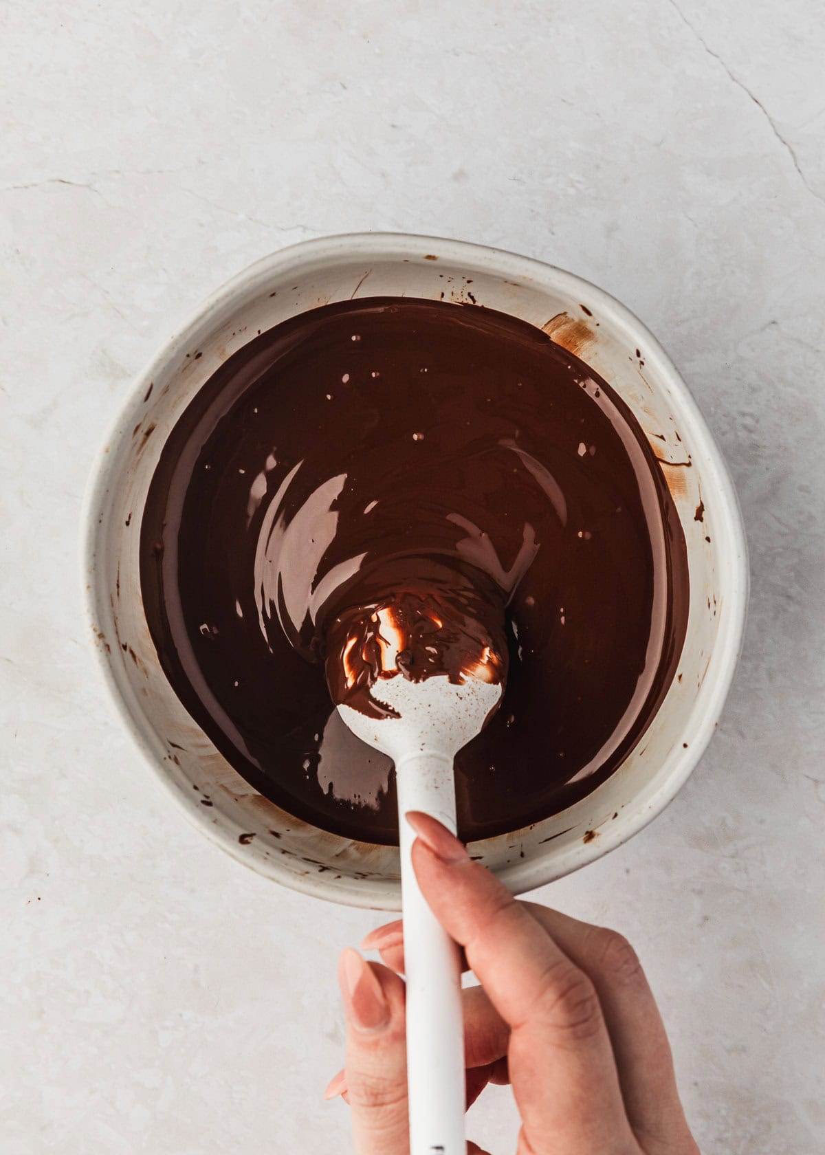 A hand using a rubber spatula to mix melted chocolate in a white bowl.