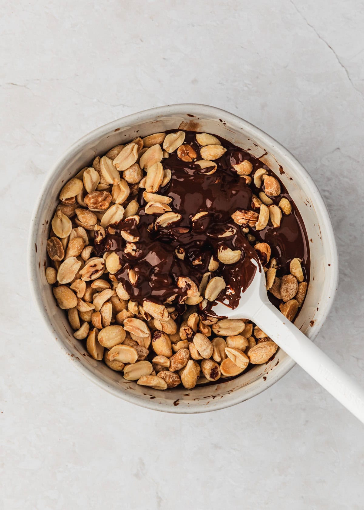 A white bowl of chocolate and peanuts for candy clusters on a white counter.