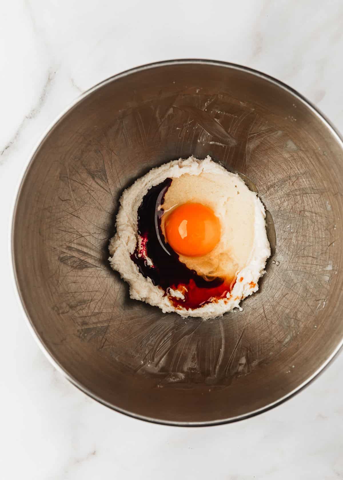 A silver bowl of butter, sugar, egg, vanilla, and red food coloring on a white counter.