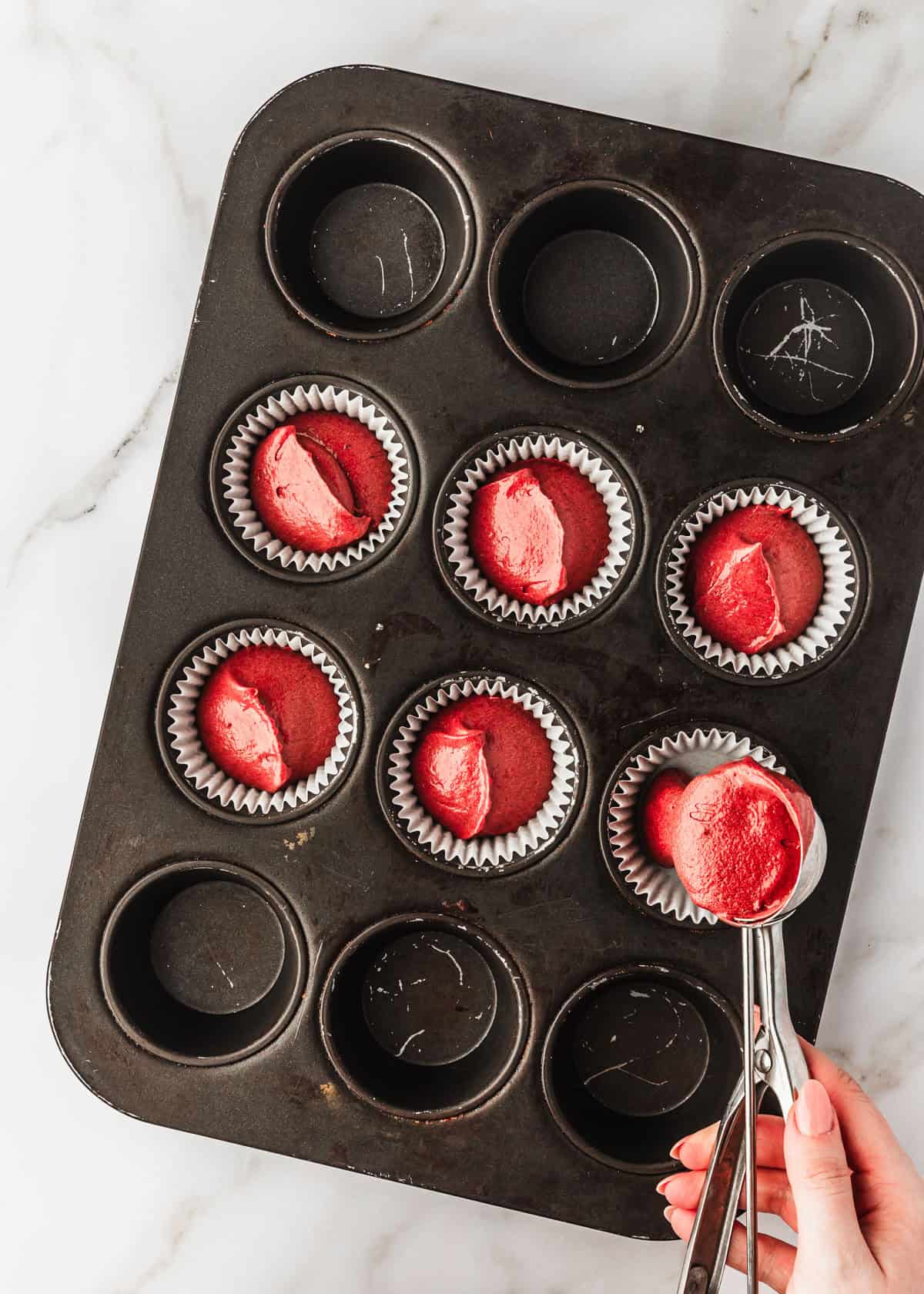 A hand using a scoop to divide batter for small batch red velvet cupcakes into a cupcake pan.