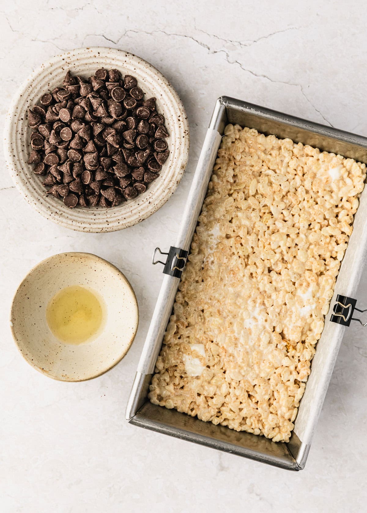 A pan of cereal bars next to white bowls of chocolate chips and oil on a white counter.