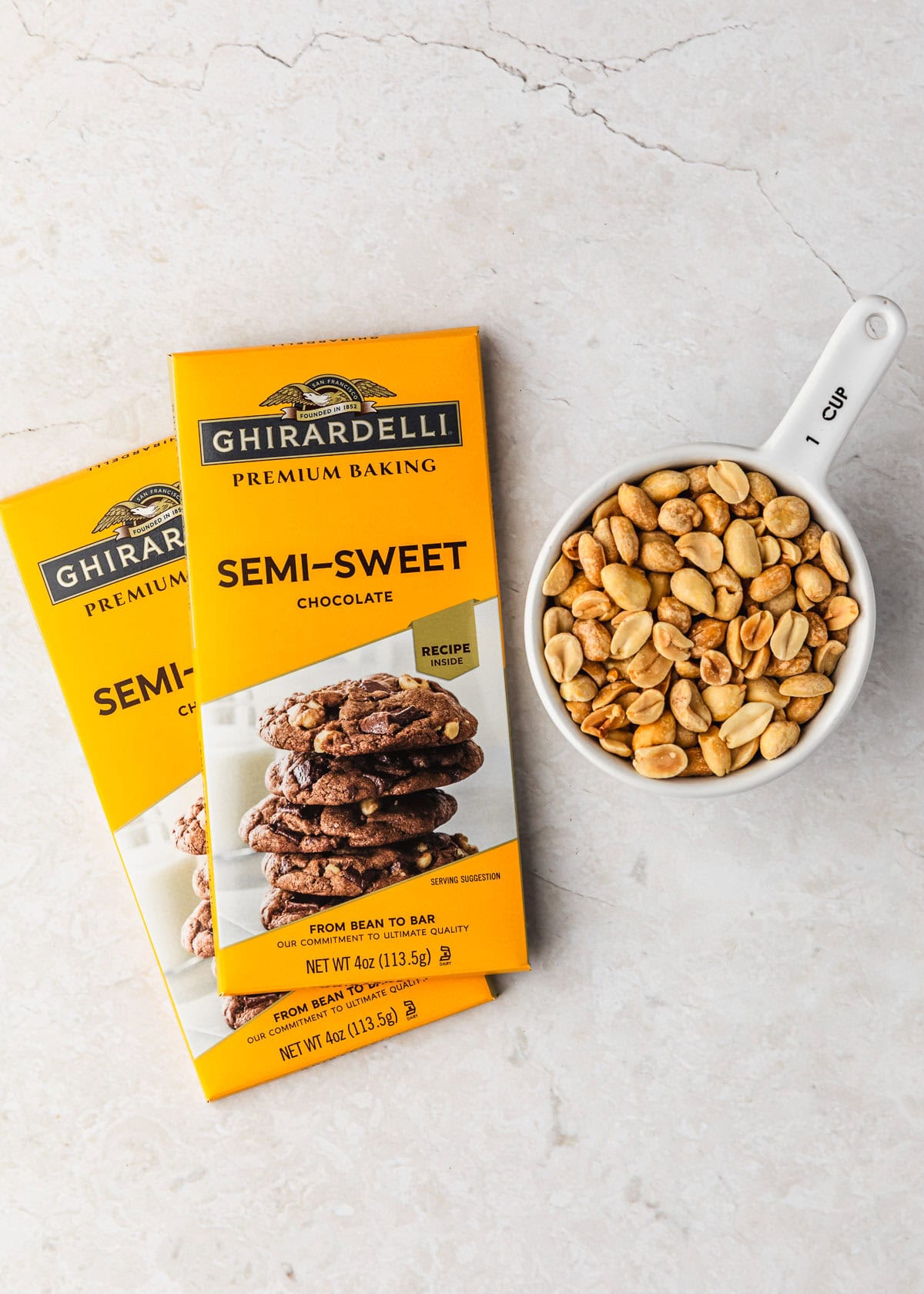 Bars of semi-sweet chocolate and a white measuring cup of peanuts on a white counter.