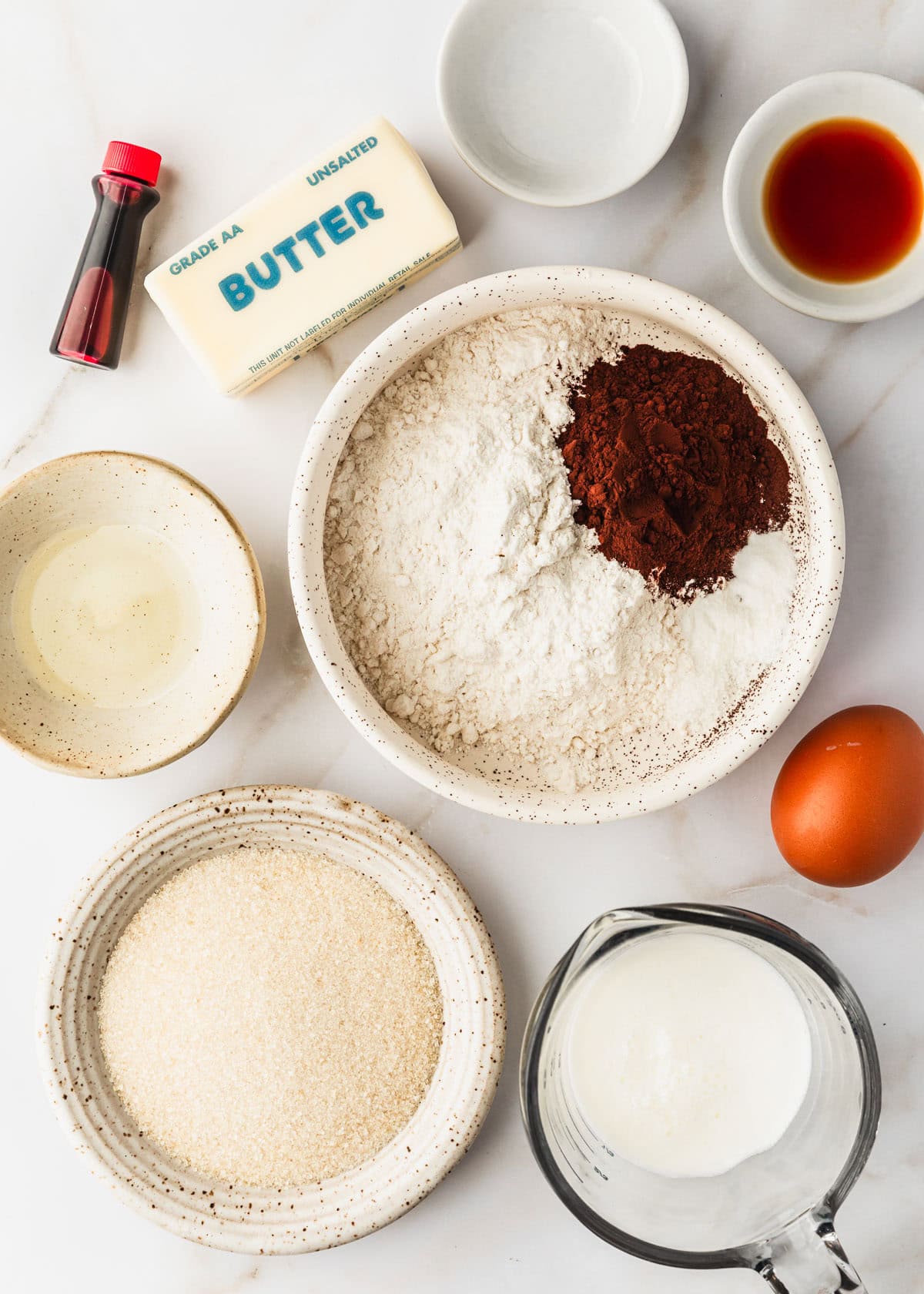 White bowls of flour, cocoa, oil, sugar, vanilla, and vinegar next to a stick of butter, egg, cup of buttermilk, and vial of red food coloring on a white counter.