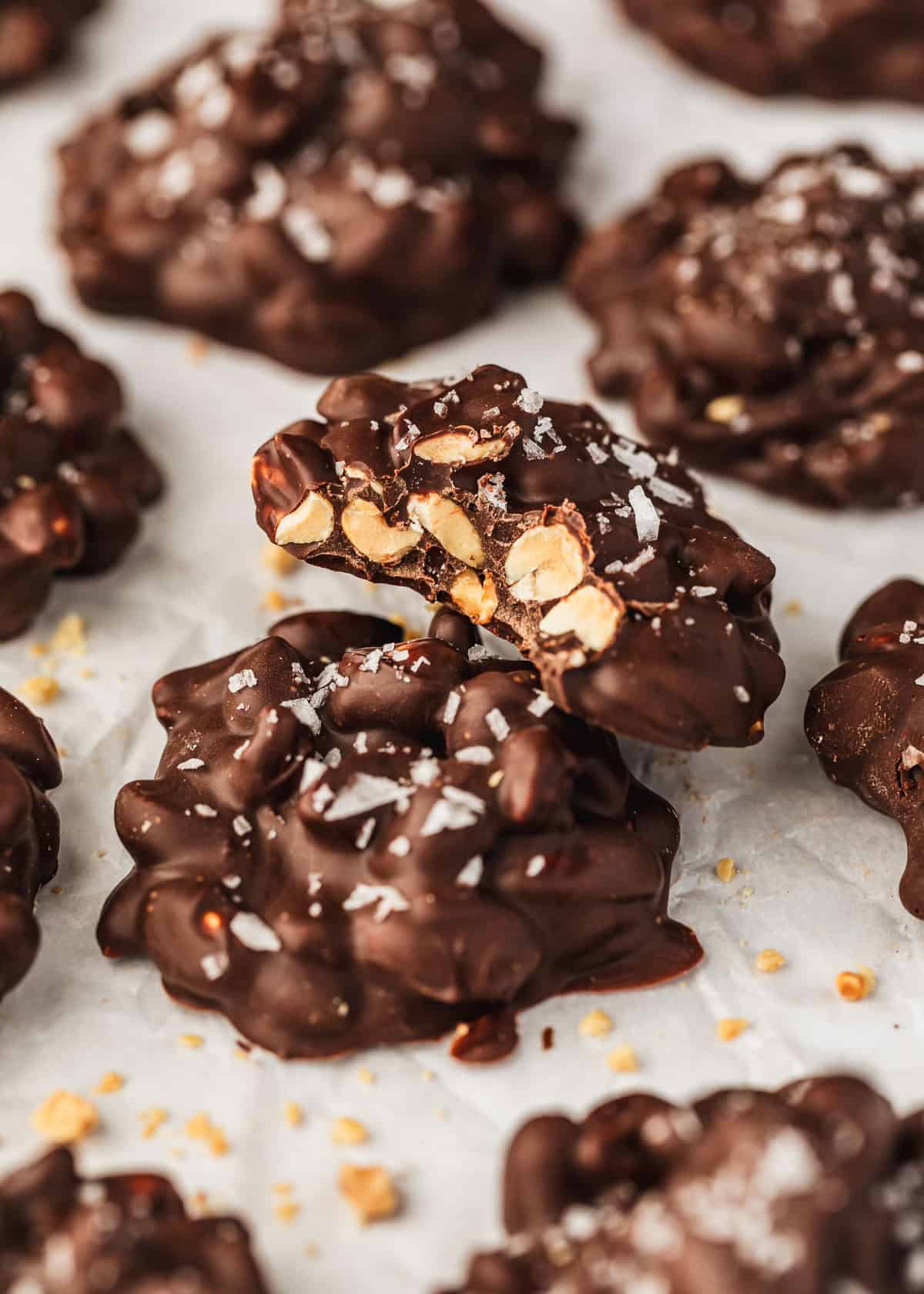 A chocolate candy leaning against another candy next to rows of chocolate peanut clusters on parchment paper.