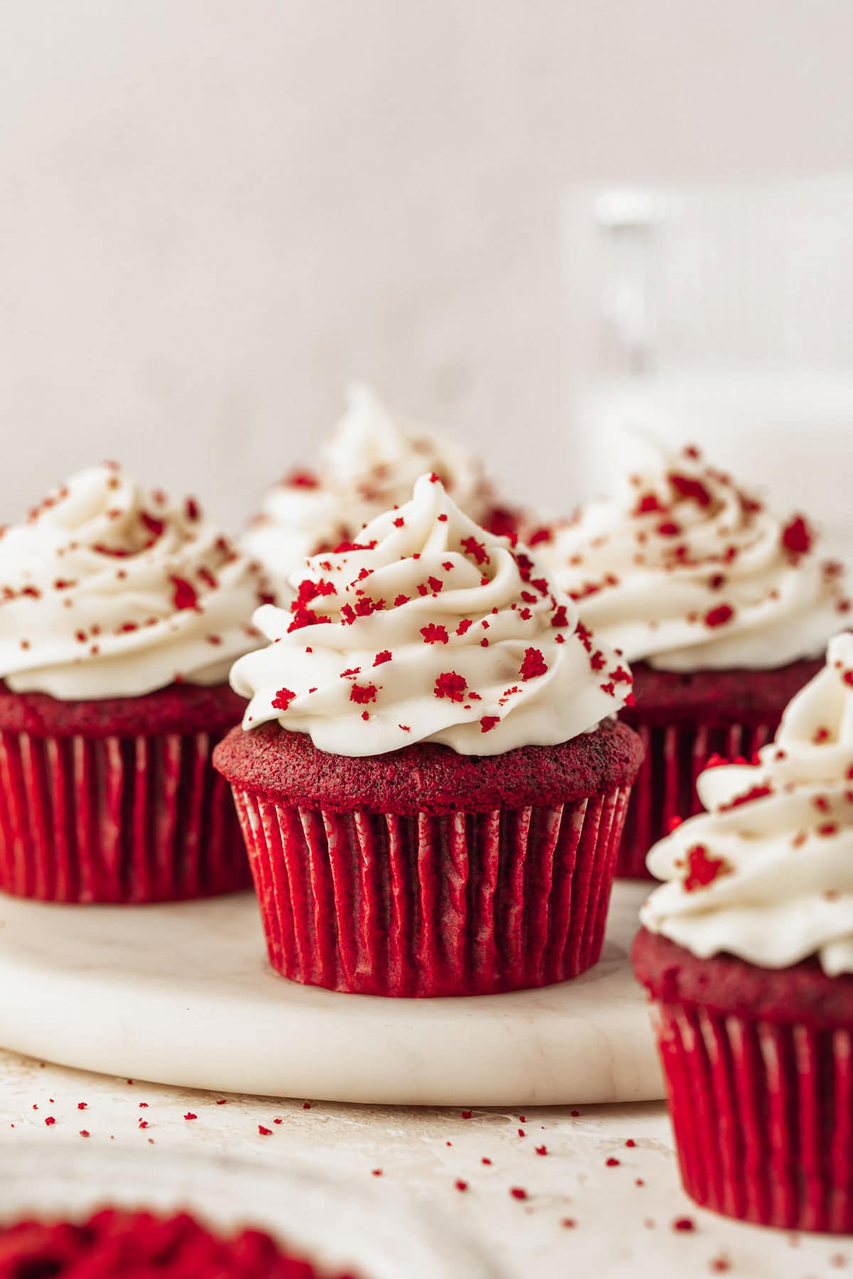 A white tray of small batch red velvet cupcakes with cream cheese frosting next to a white bowl of red velvet crumbs on a beige counter.