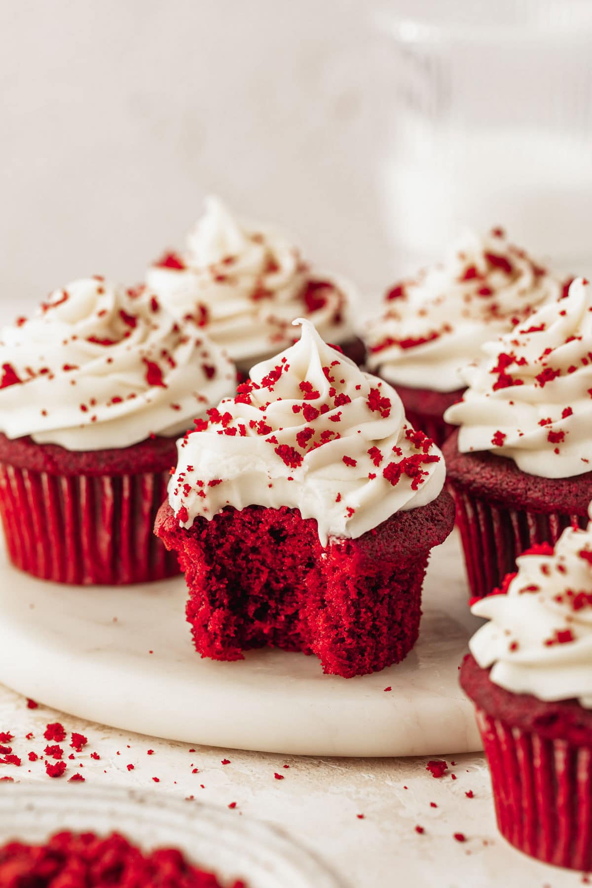 A cupcake with a bite taken out of it next to rows of small batch red velvet cupcakes on a white platter. The platter is next to a glass of milk and white bowl of red velvet crumbs with a beige background.