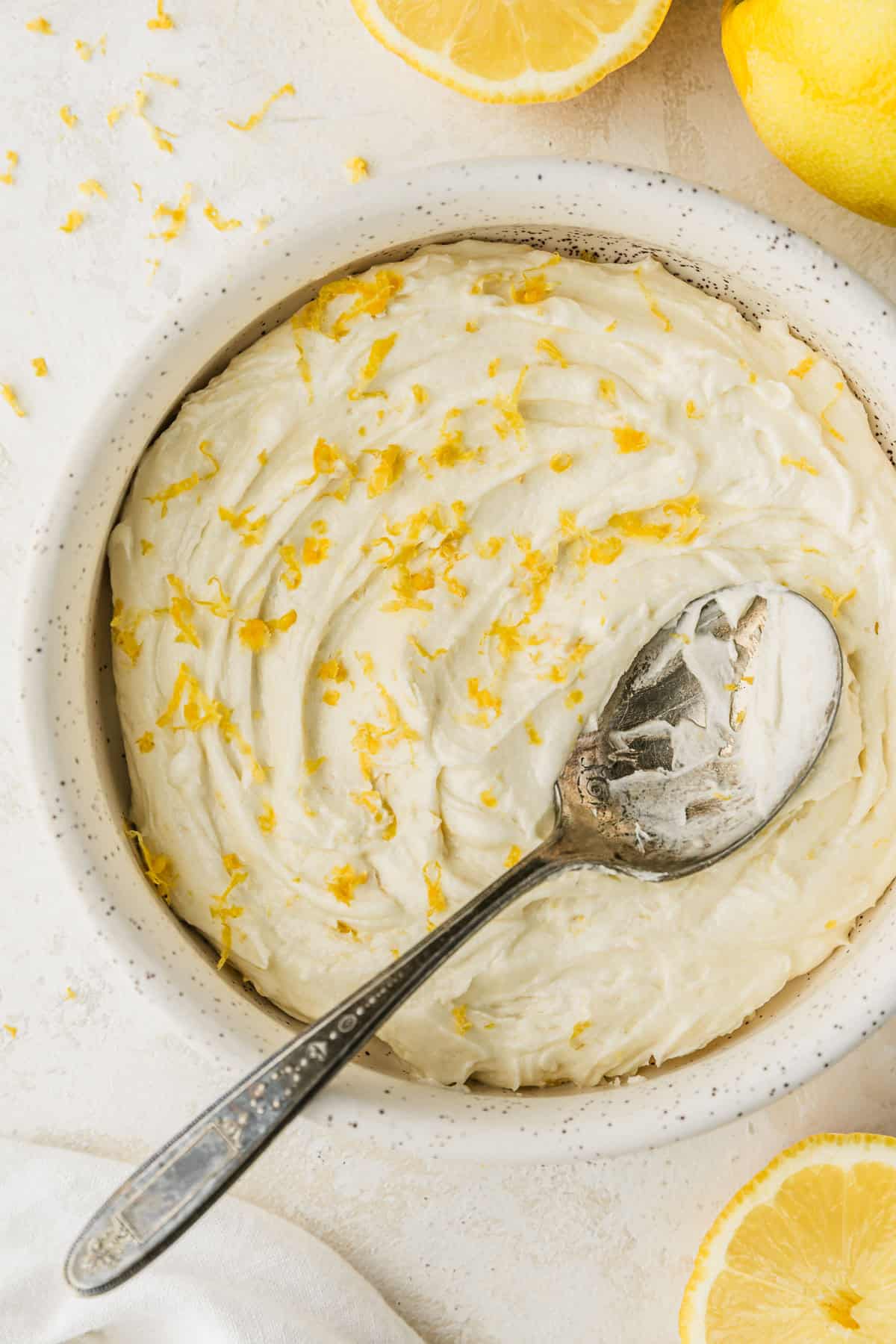 A white bowl of small batch lemon buttercream on a white counter next to lemons and a white linen.