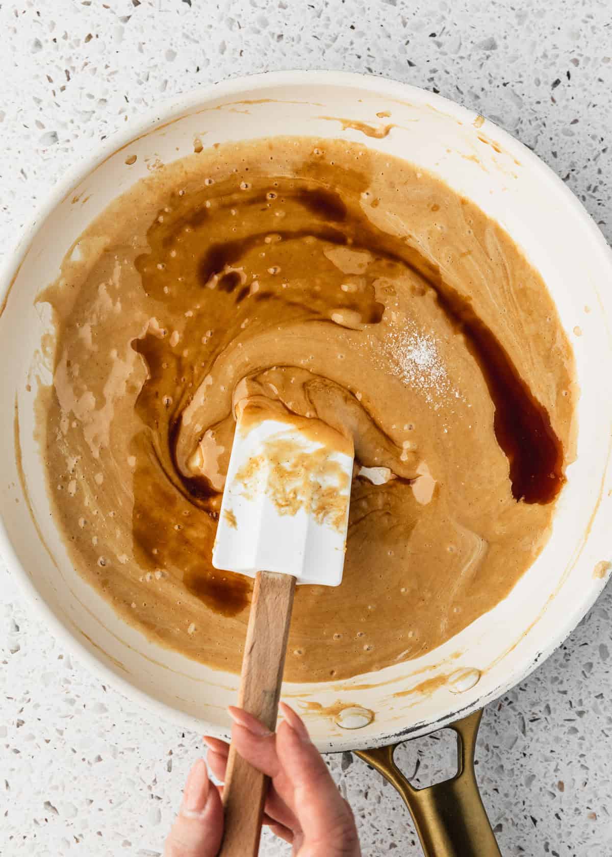 A hand using a rubber spatula to mix vanilla and salt into a white pan of melted marshmallows and peanut butter.
