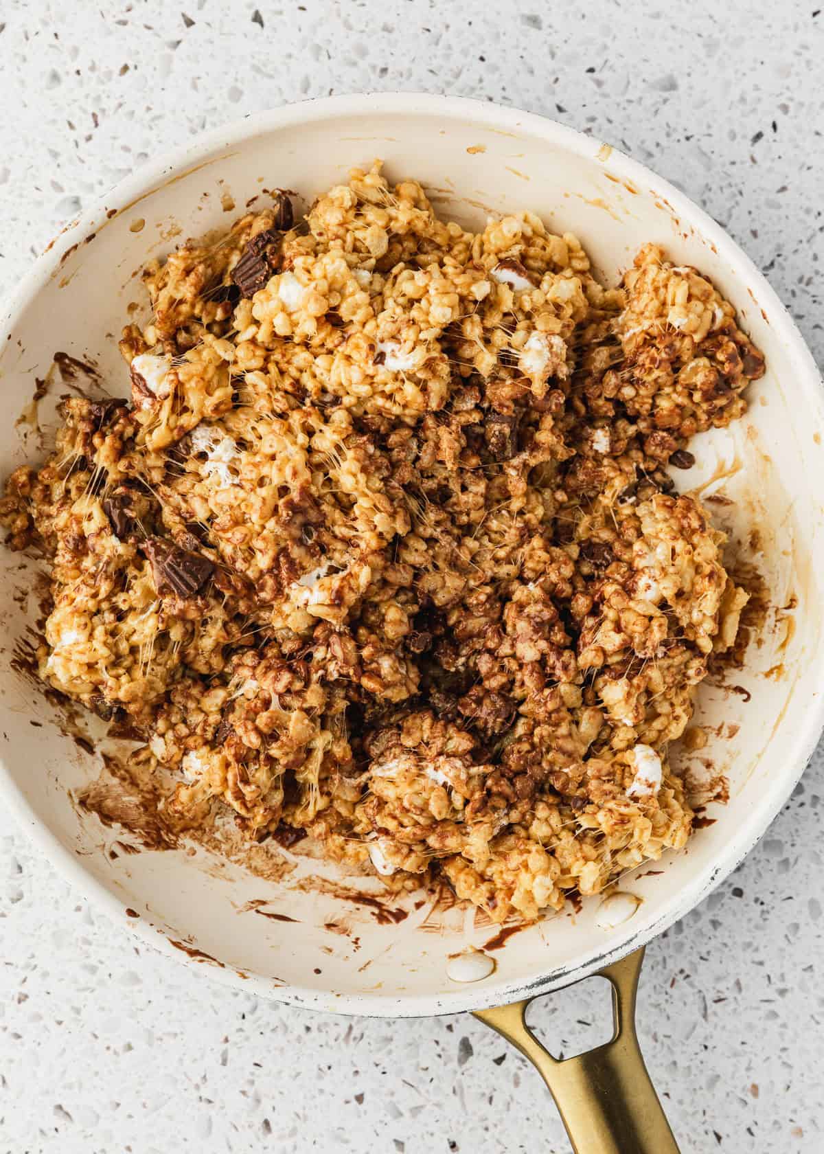 A white pan of rice krispies, chocolate peanut butter cups, peanut butter, and marshmallows on a white counter.