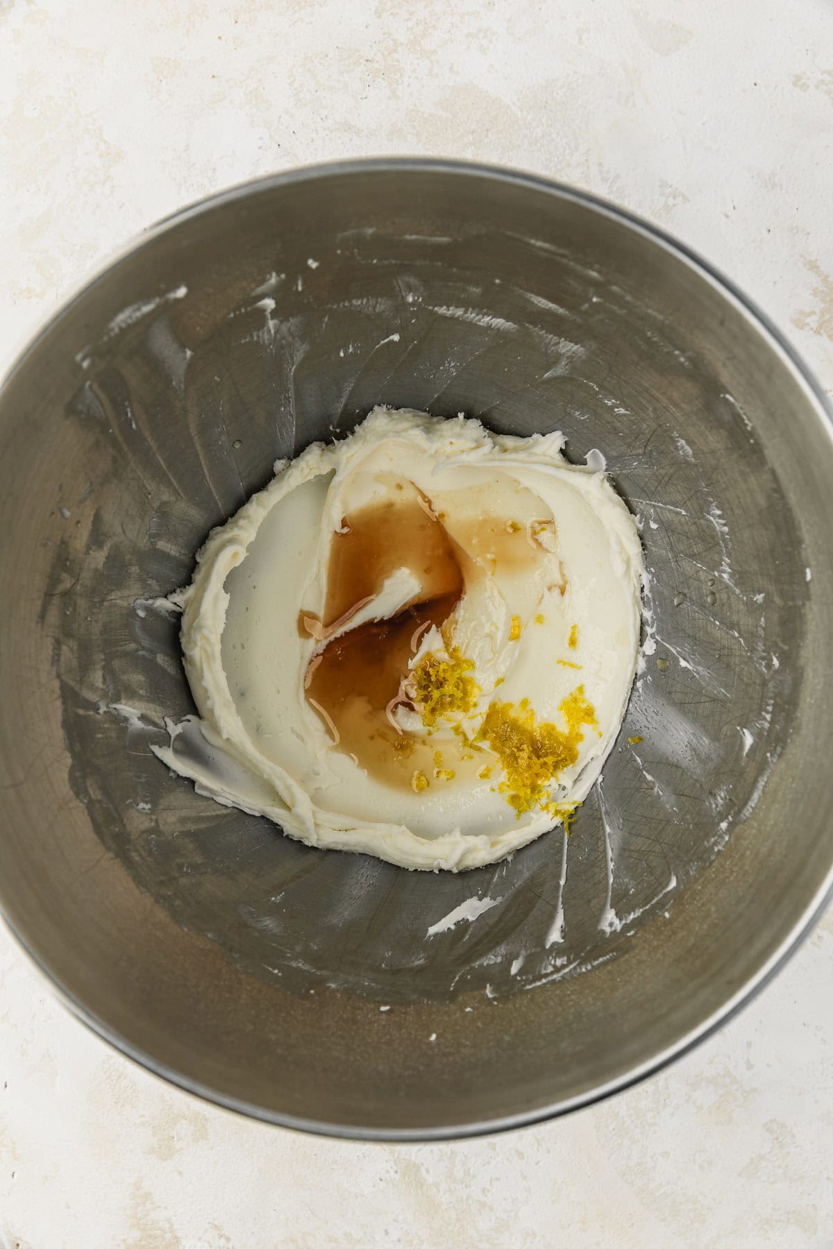 A silver bowl of frosting with vanilla extract and lemon zest on a white counter.
