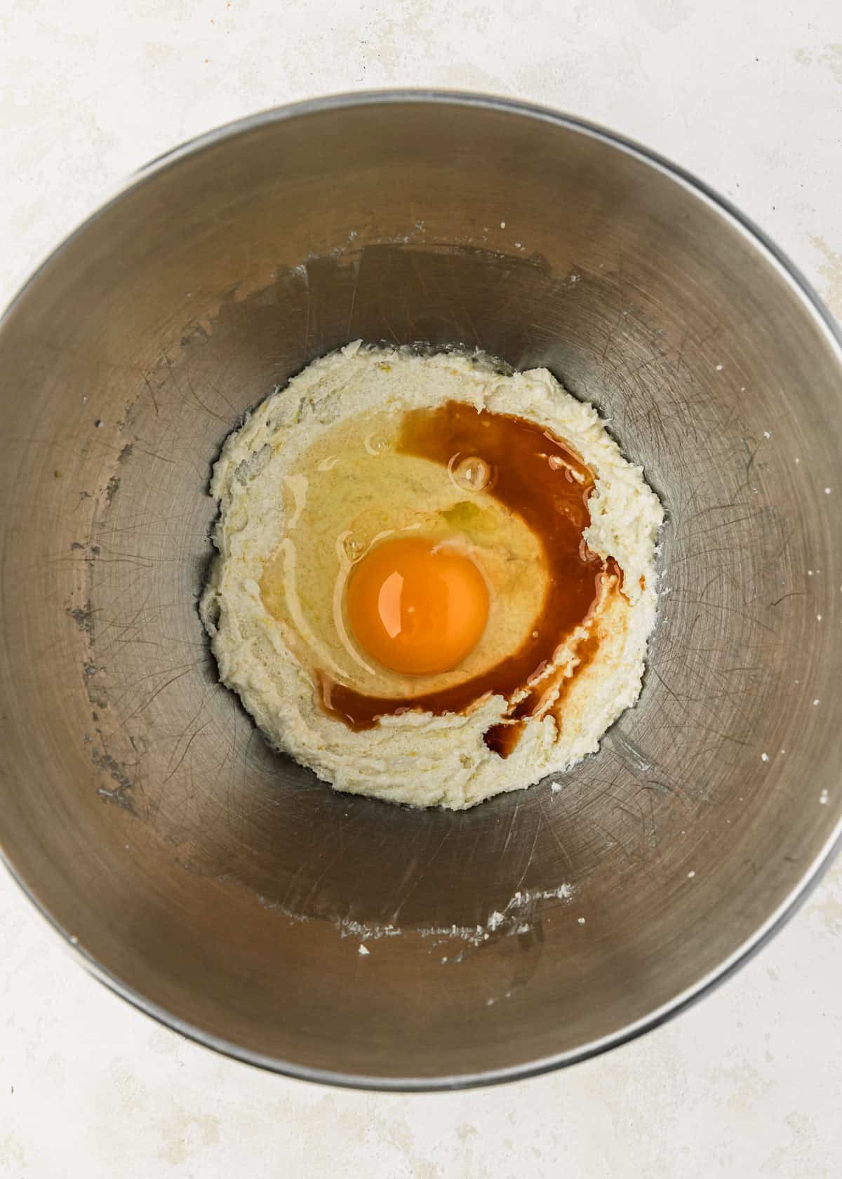 A silver bowl of creamed butter and sugar, vanilla, and an egg on a tan counter.