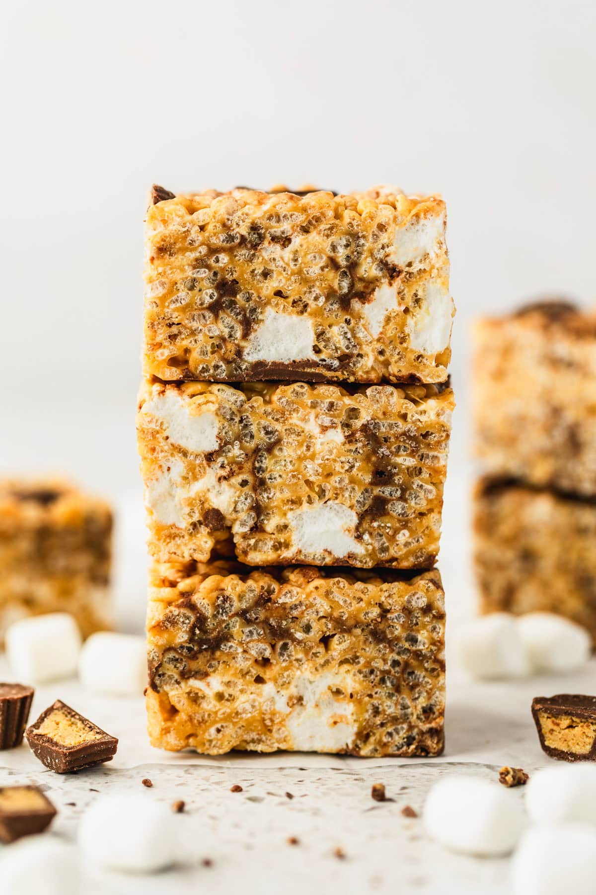 A stack of small batch chocolate peanut butter rice krispie treats next to more treats, marshmallows, and peanut butter cups on a white counter.