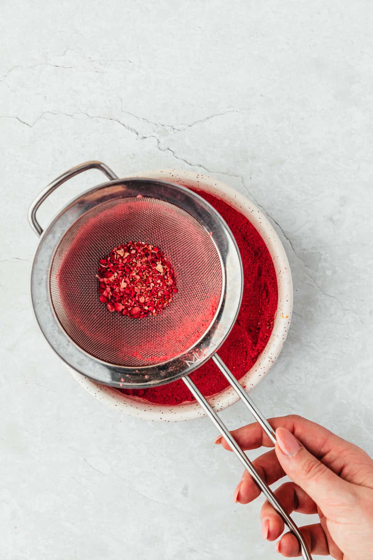 A hand using a sieve to sift small batch strawberry powder into a white bowl.